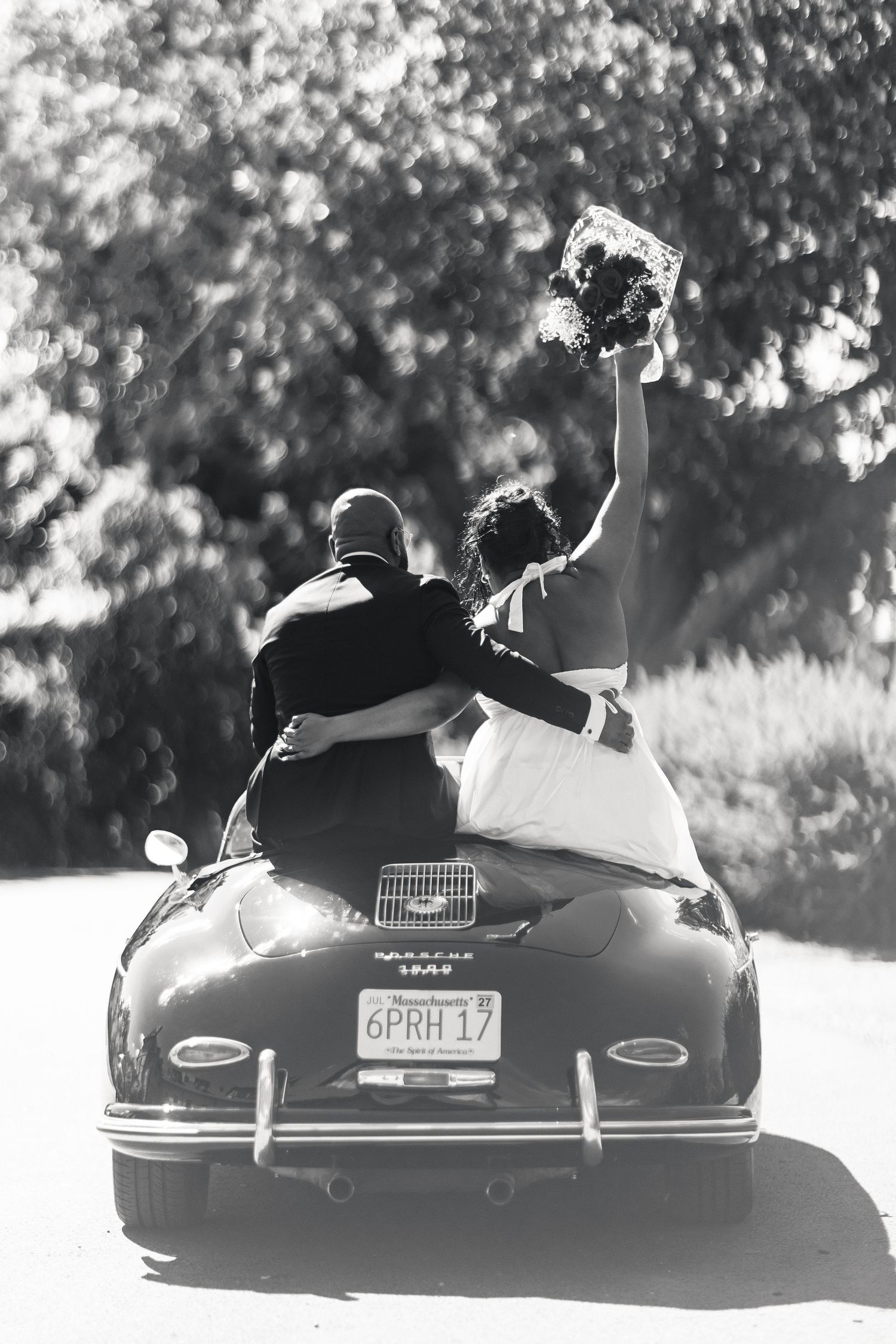 Black and white photo of newlyweds sitting on vintage Porsche as bride raises bouquet in celebration.