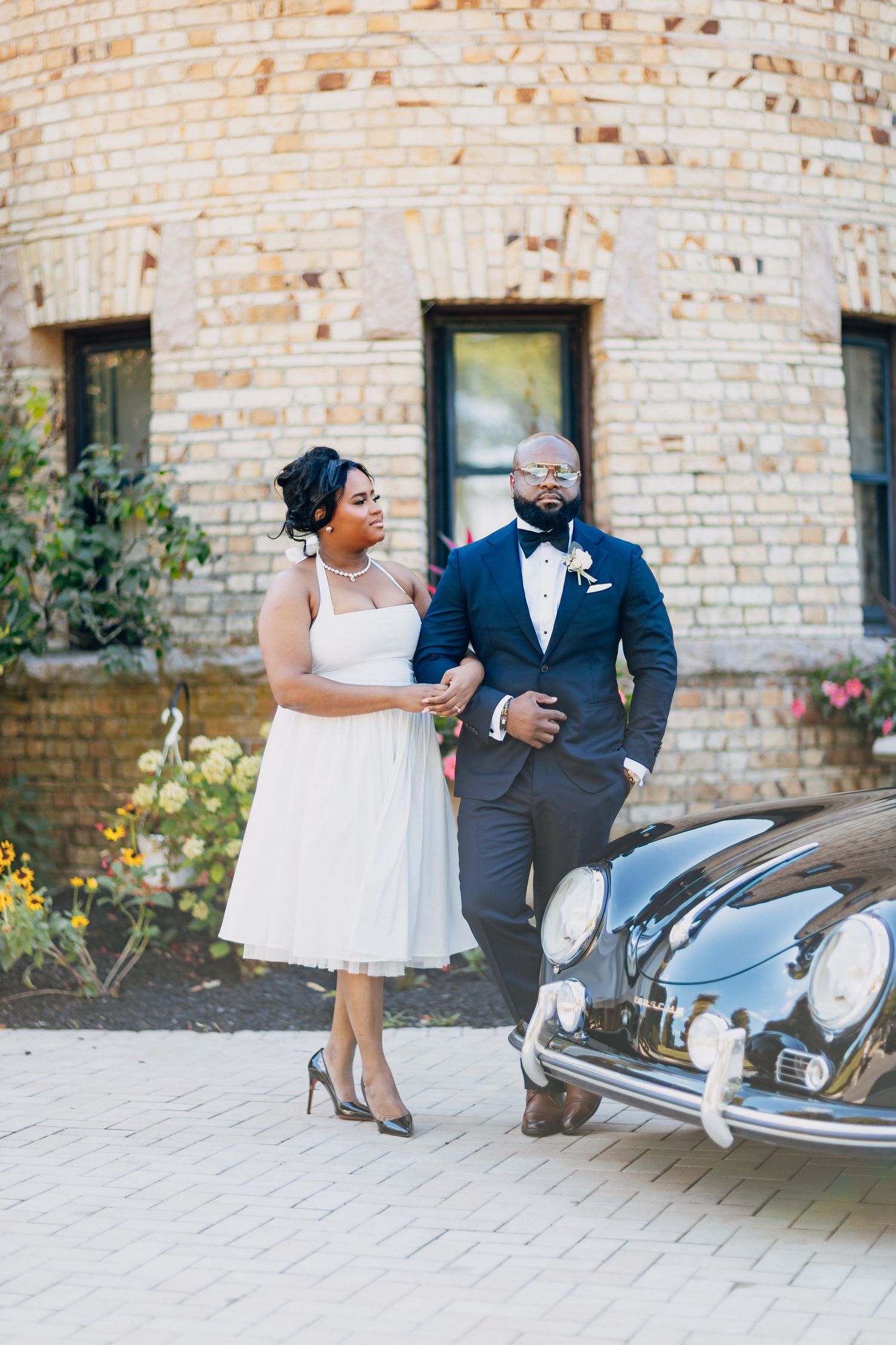 Bride in white midi dress and groom in navy tuxedo pose beside a vintage black convertible outside a brick building.