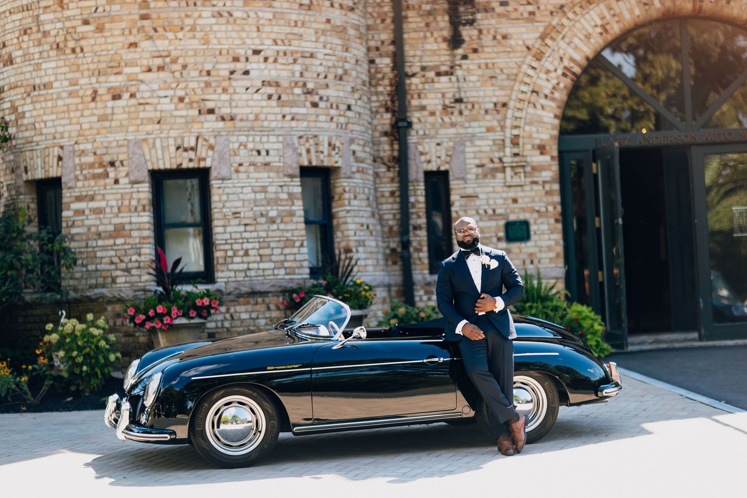 Dapper groom in navy tuxedo leans against classic black Porsche 356 outside brick wedding venue.