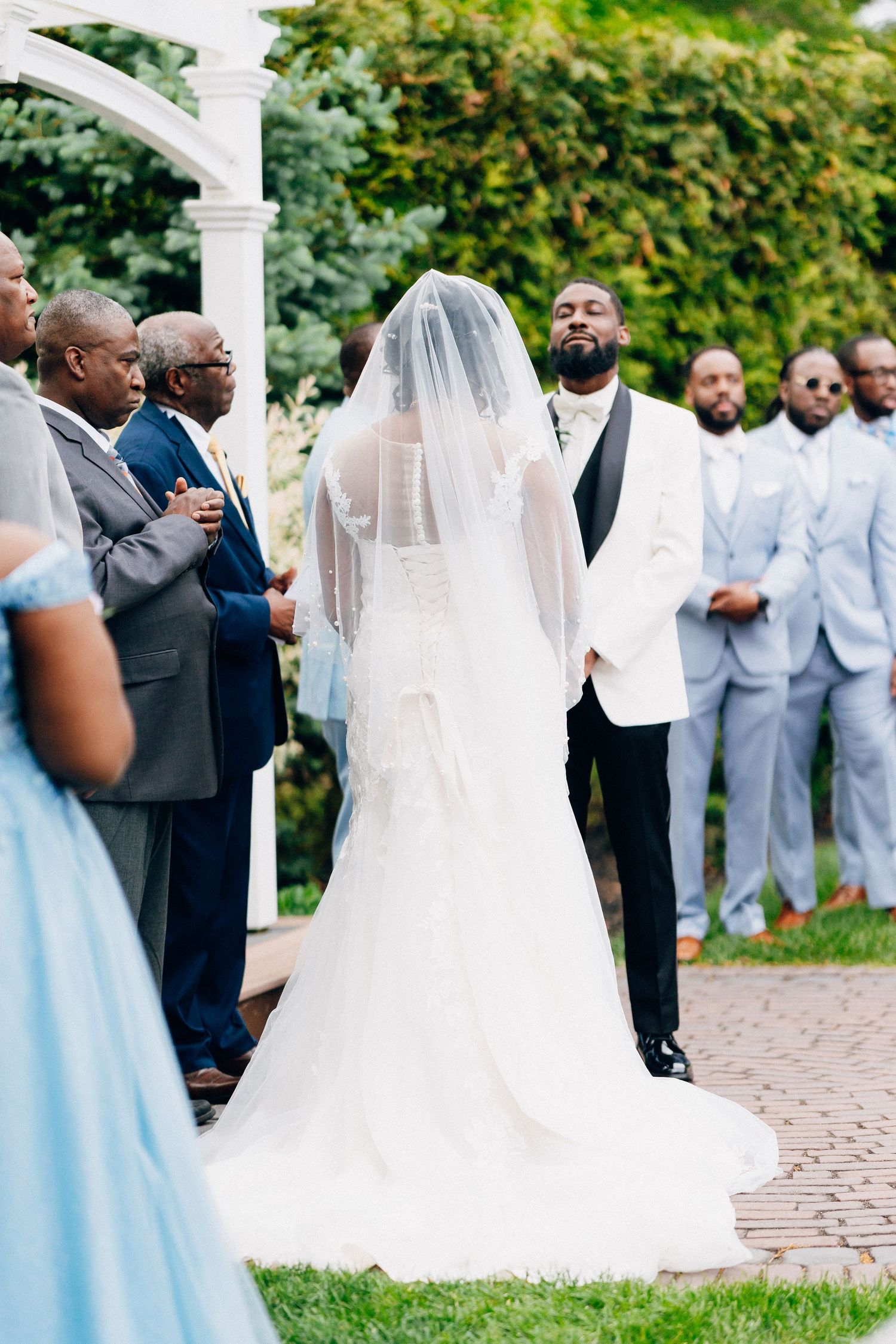 Groom in white tuxedo jacket emotional at altar as bride in white gown and veil approaches during outdoor wedding ceremony.