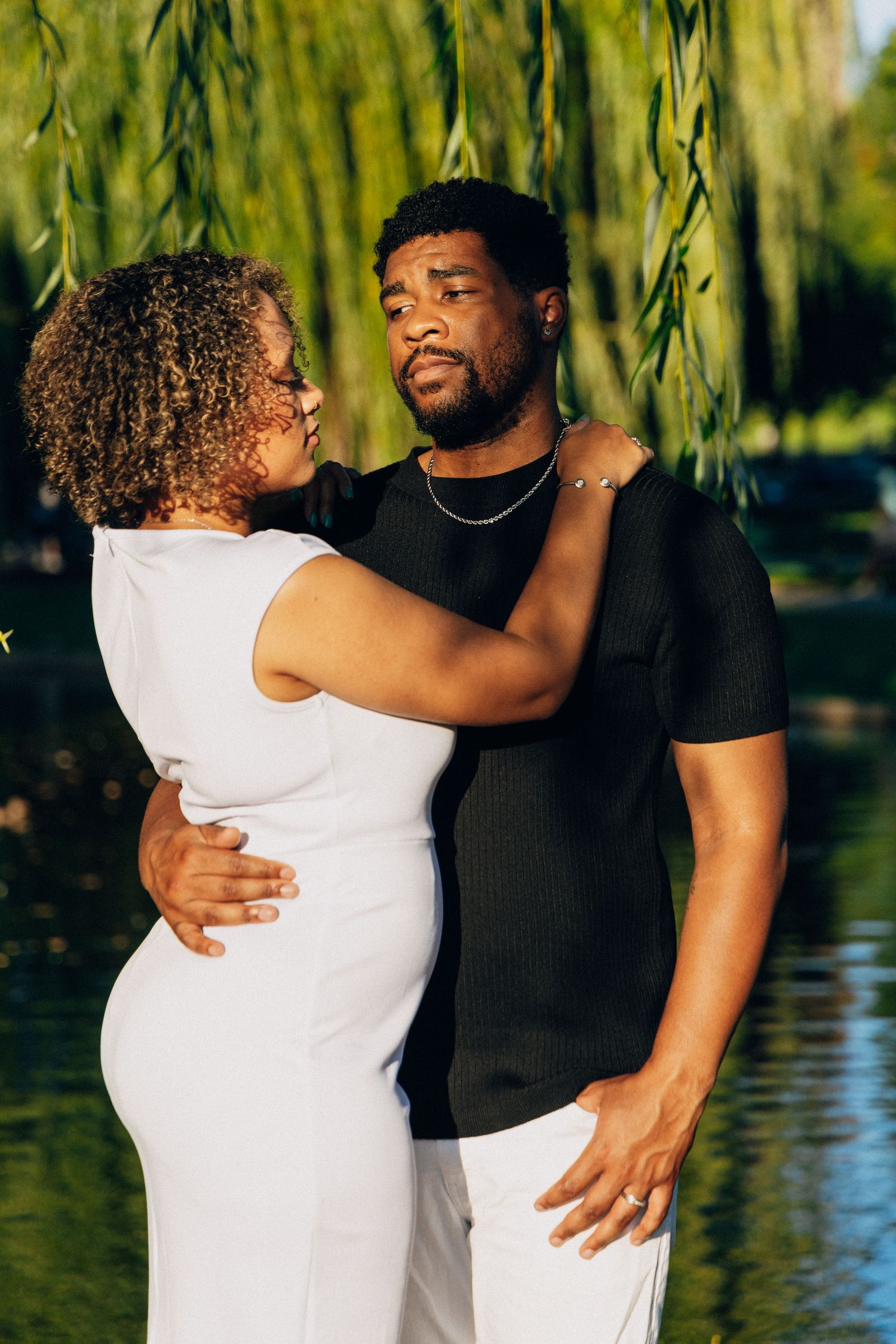 A couple embraces near a willow tree by a lake, woman in white dress and man in black shirt.