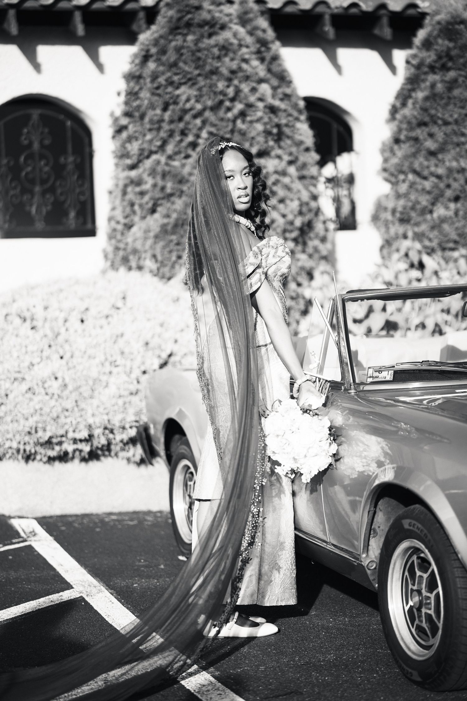 Black and white photo of bride in teal gown with long veil beside vintage convertible car.