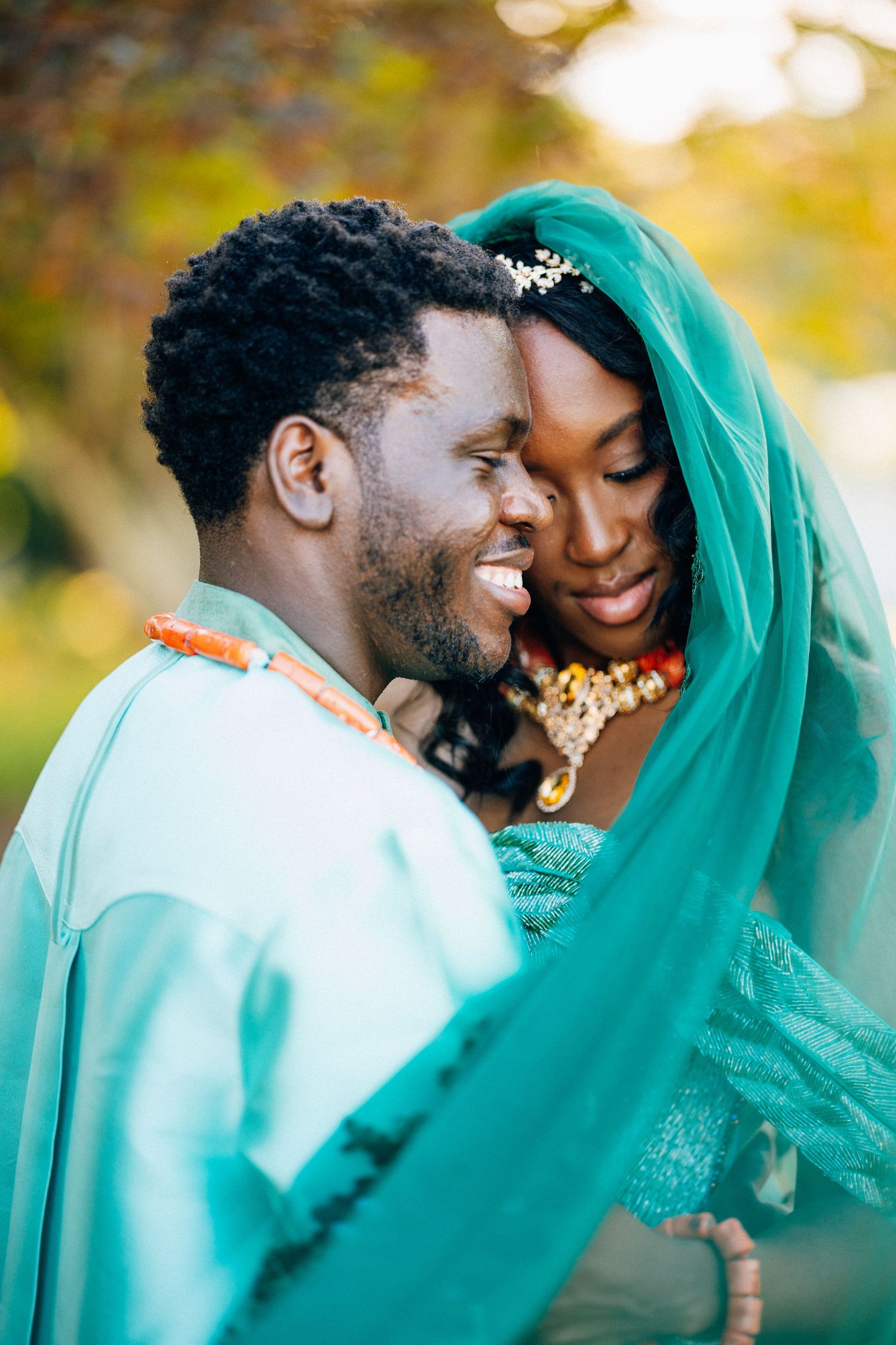 Smiling groom gazes at bride during traditional Nigerian wedding ceremony, both adorned in teal attire and coral beads.