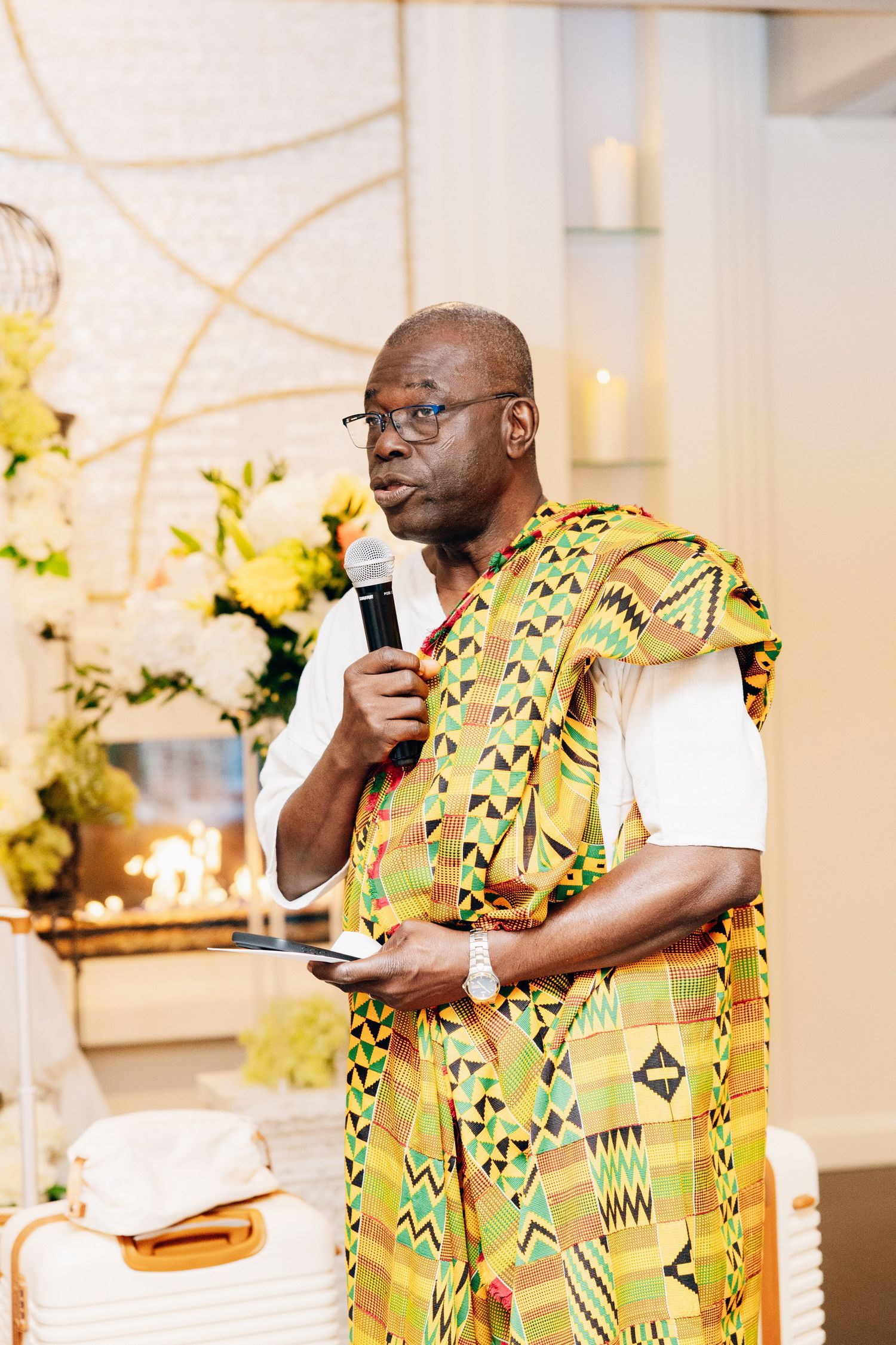 Elder man in colorful Kente cloth speaks at wedding ceremony holding microphone.