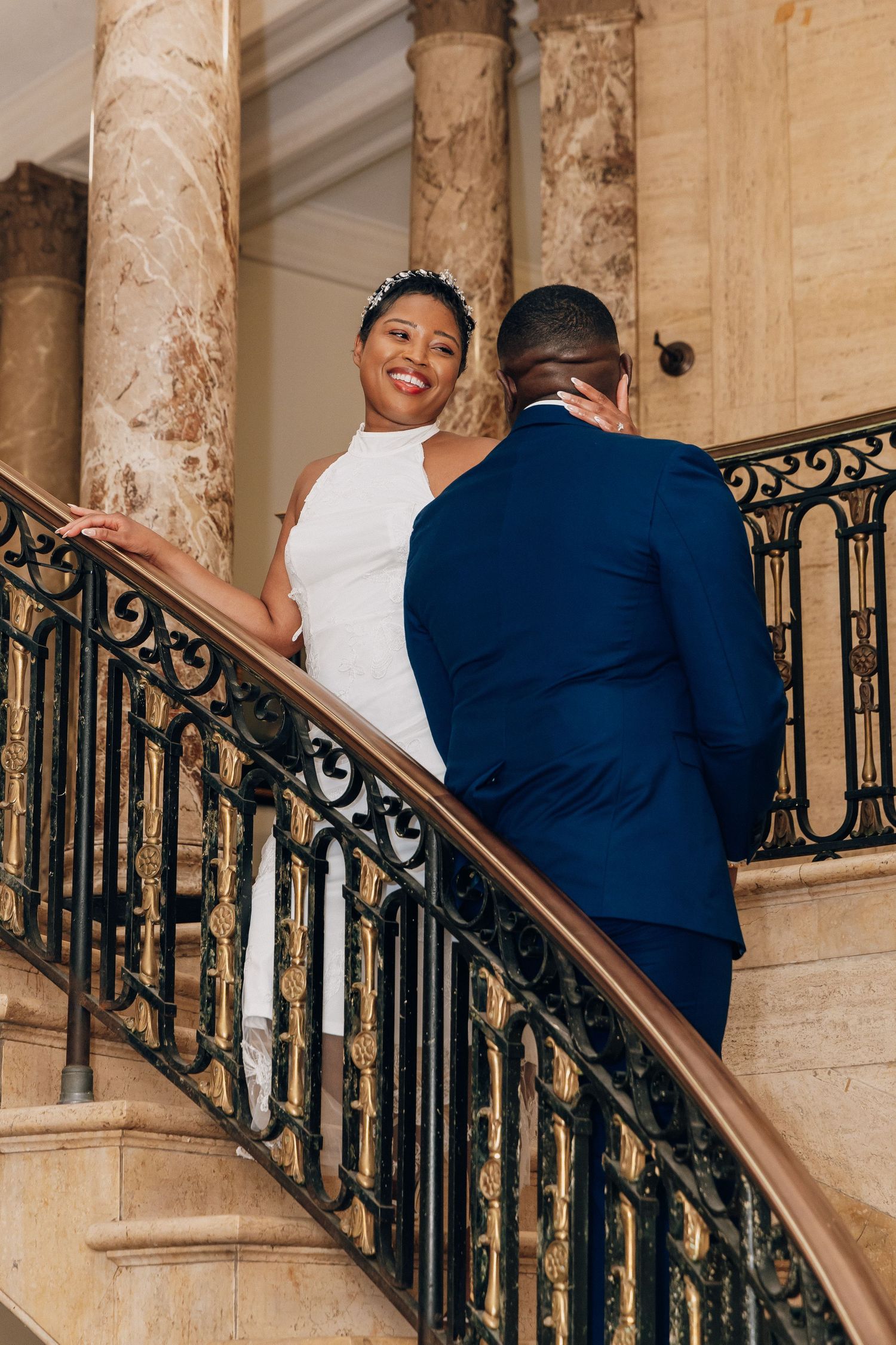 Bride in white dress and groom in navy suit share a romantic moment on an ornate staircase.