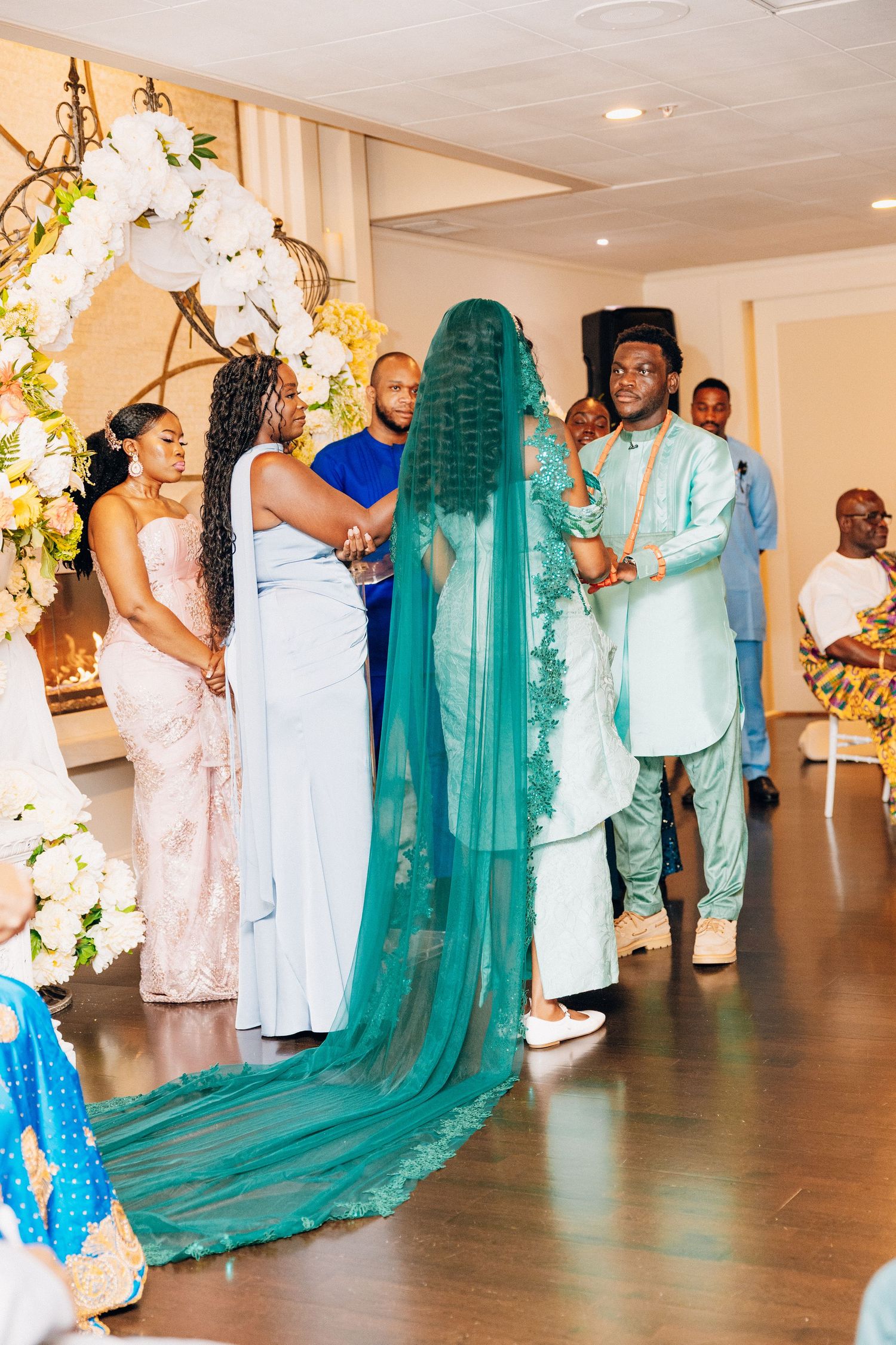 Bride in teal gown with long veil stands with groom during multicultural wedding ceremony with floral decor.
