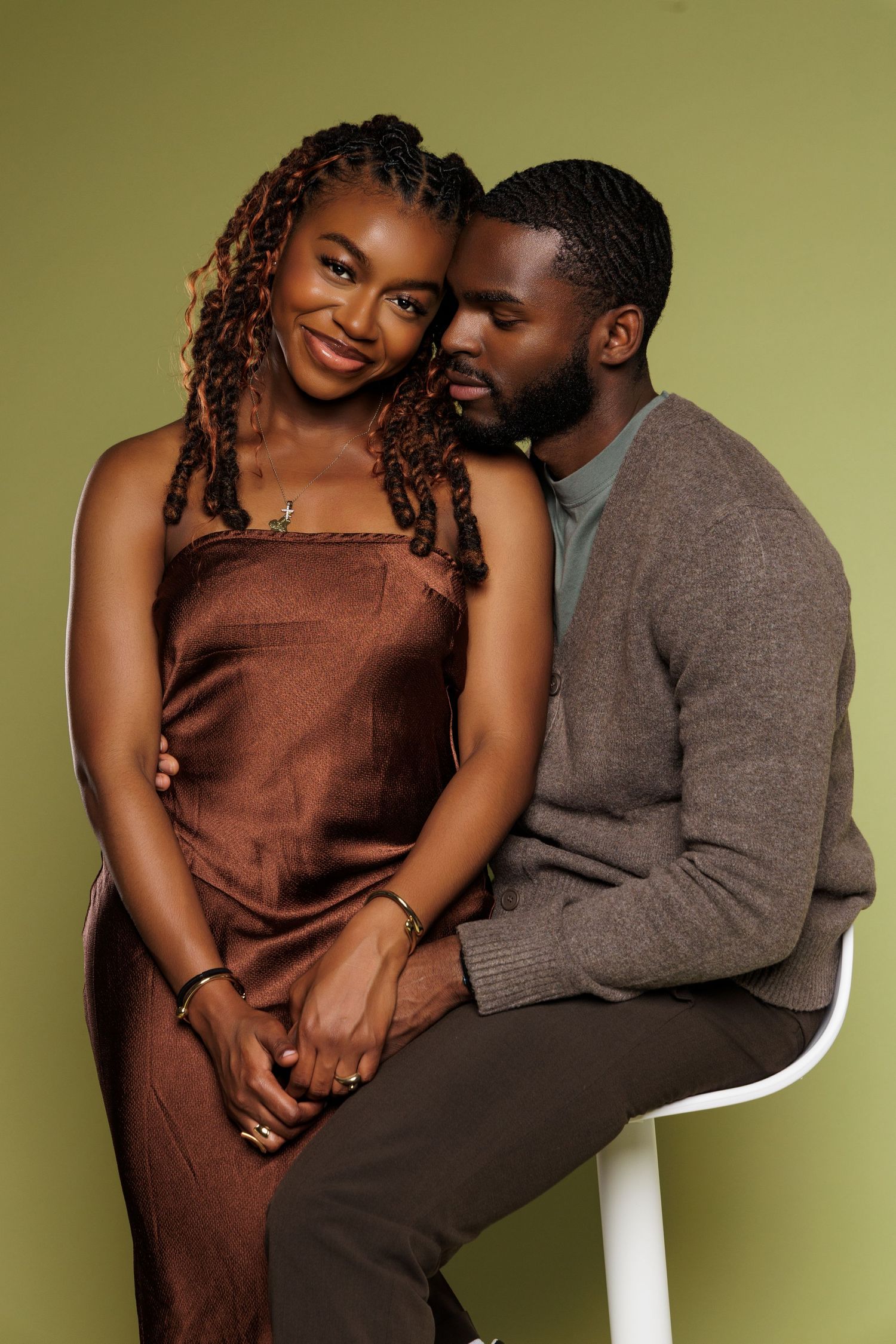 A stylish Black couple poses together in a studio, wearing earth-tone outfits against an olive green background.