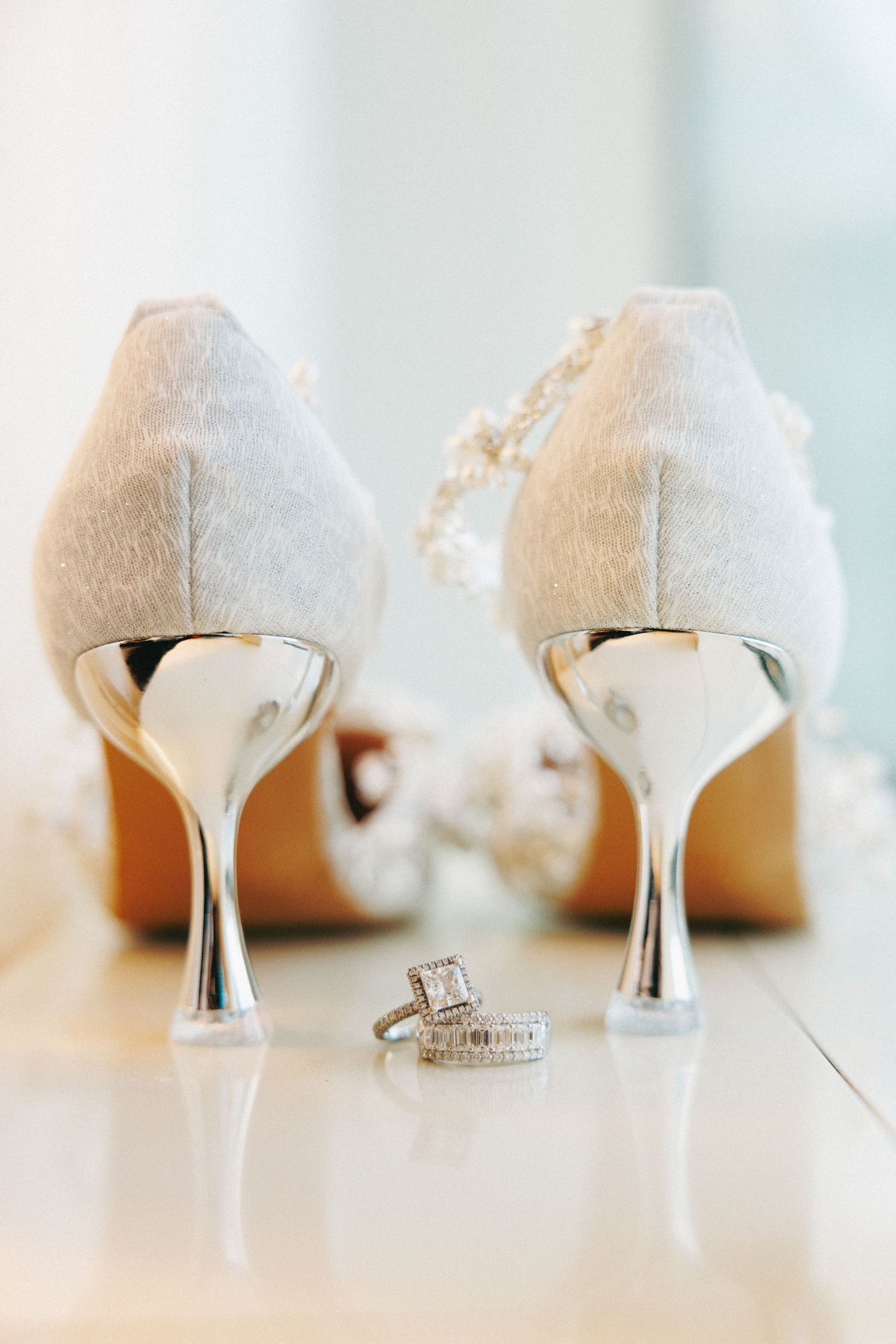 Elegant white lace bridal heels with silver stilettos and pearl ankle straps displayed with diamond wedding rings on a reflective surface.