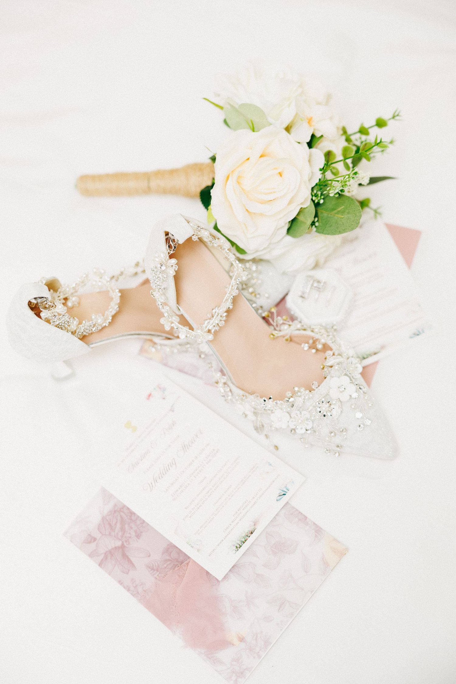 Elegant bridal heels adorned with pearls and crystals beside a white rose bouquet and wedding stationery on white fabric.