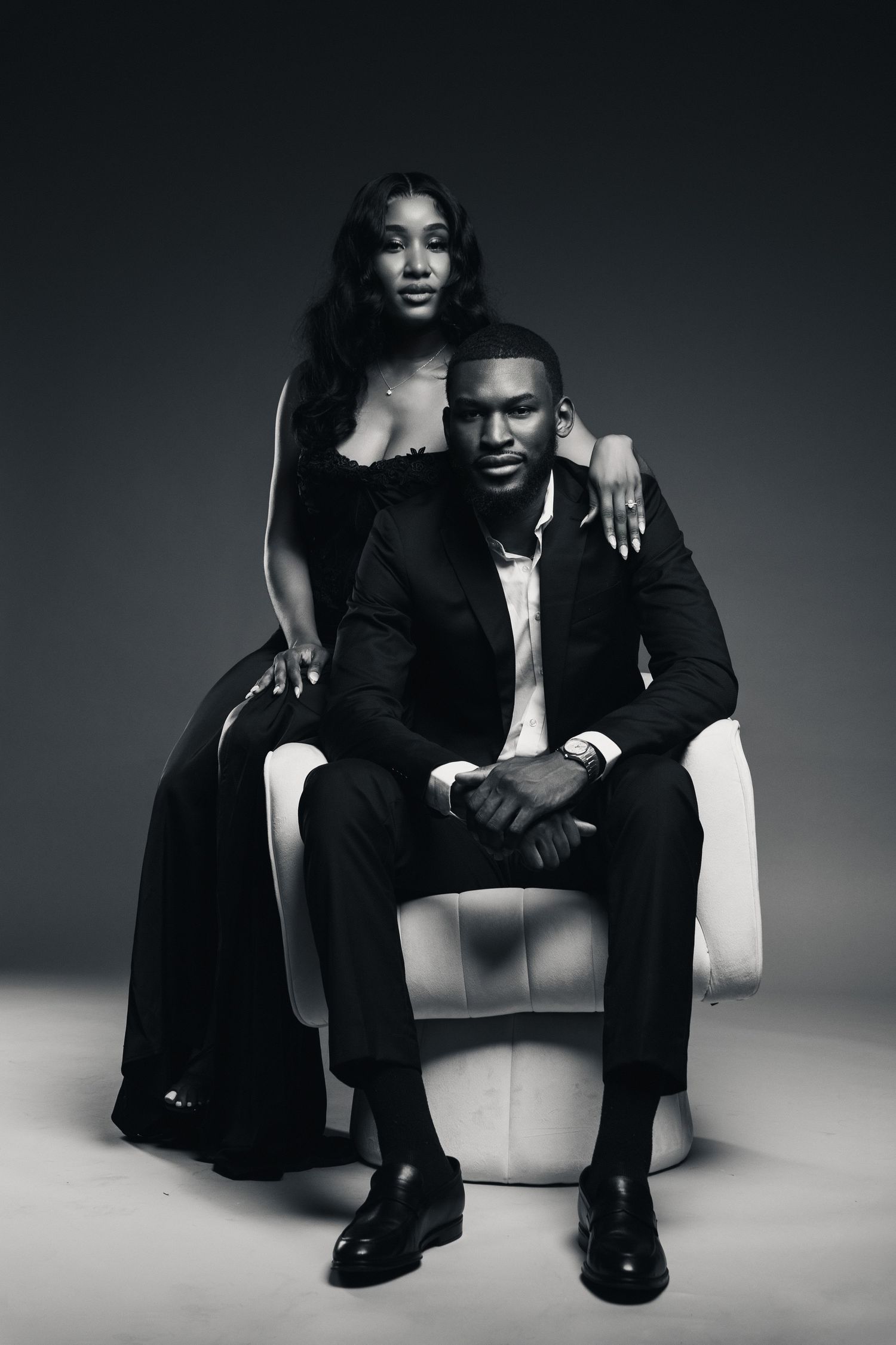 Elegant black and white portrait of a couple in formal attire, man seated in chair, woman standing behind him.