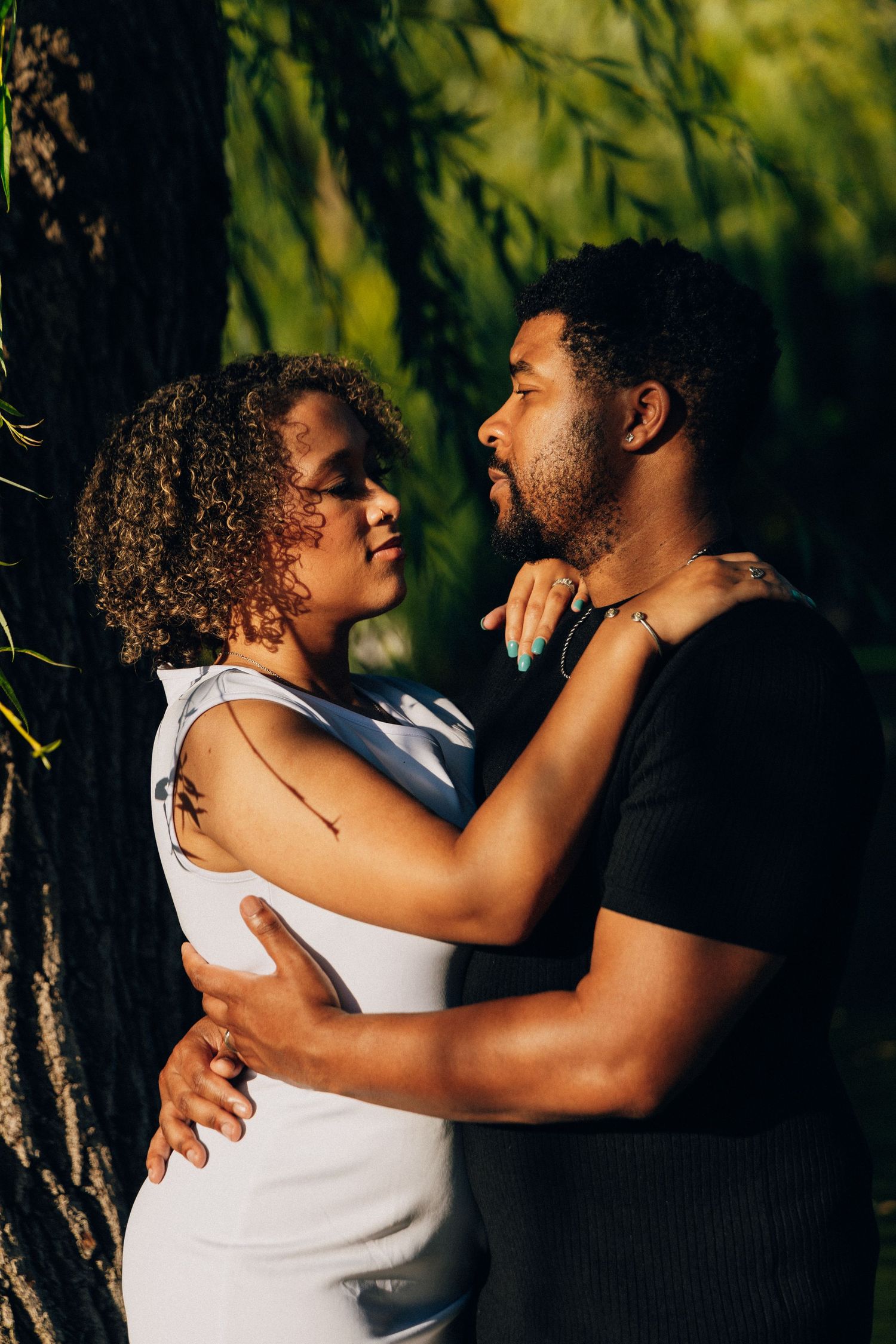 A couple shares an intimate moment face-to-face beneath a weeping willow tree in warm light.