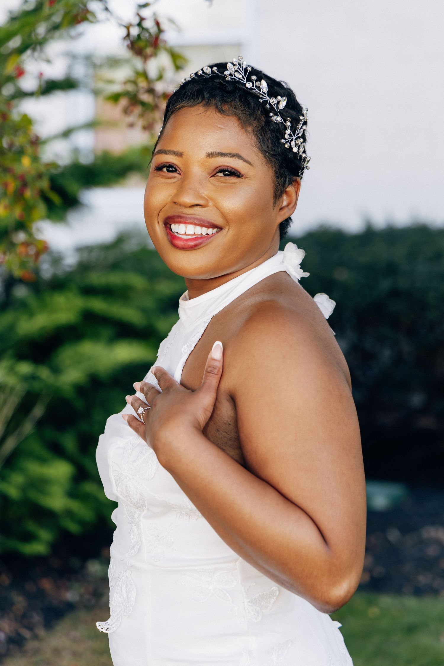 Smiling Black bride in white halter gown and crystal hair vine poses outdoors.