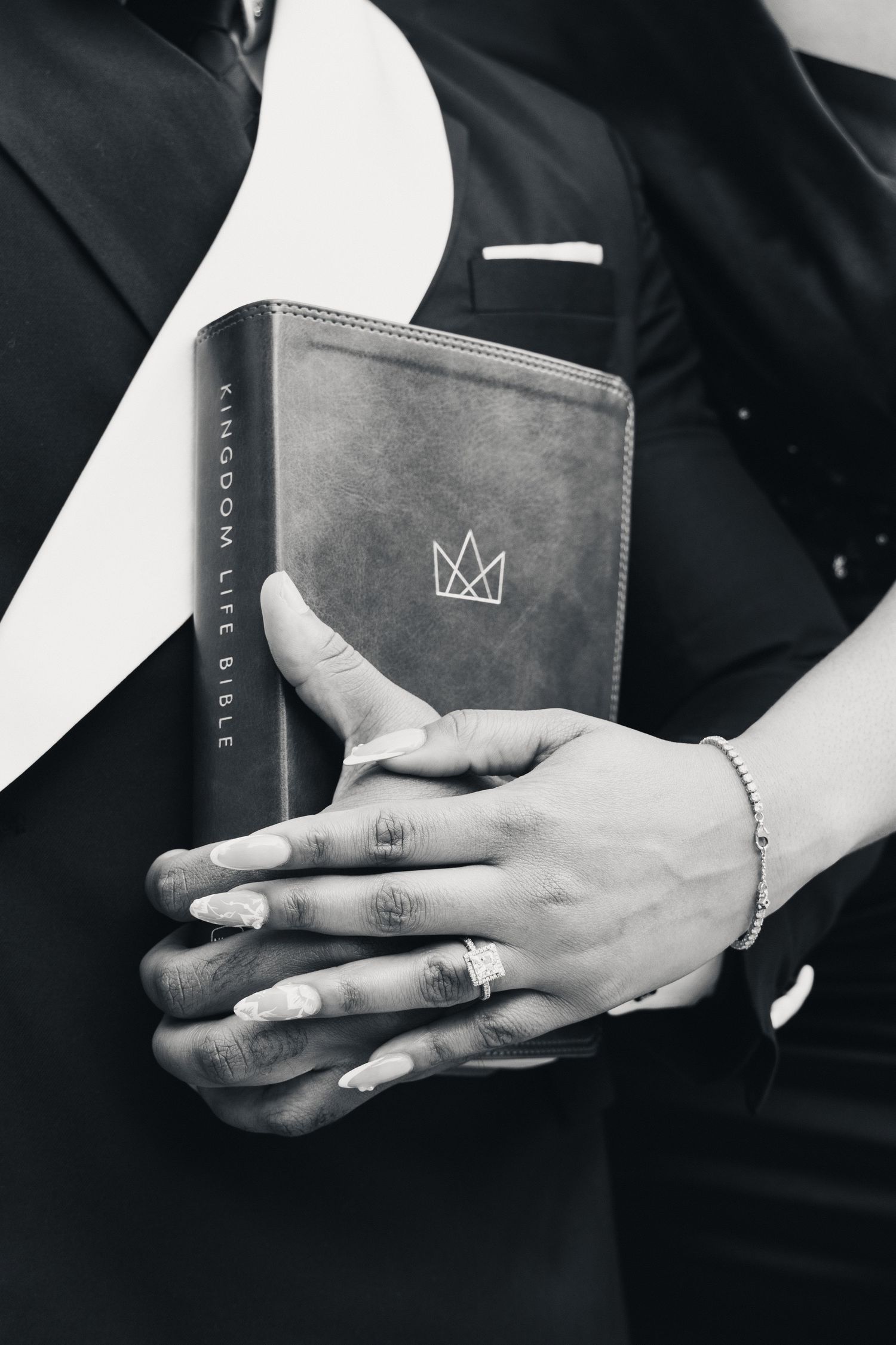 Black and white photo of hands holding a Kingdom Life Bible, with a diamond ring and bracelet visible.