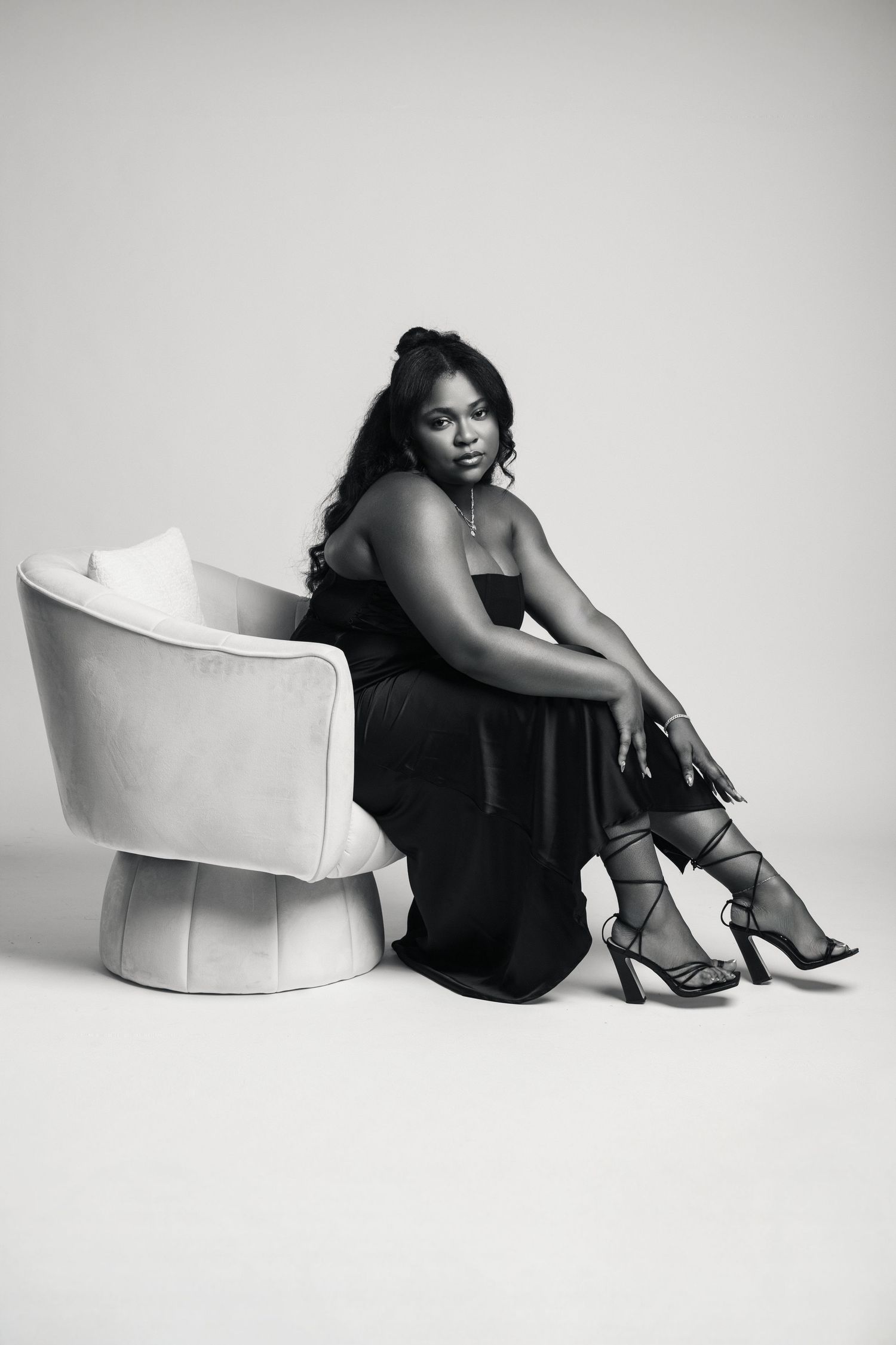 Black and white photo of a woman in an elegant black gown seated gracefully on a modern round chair.