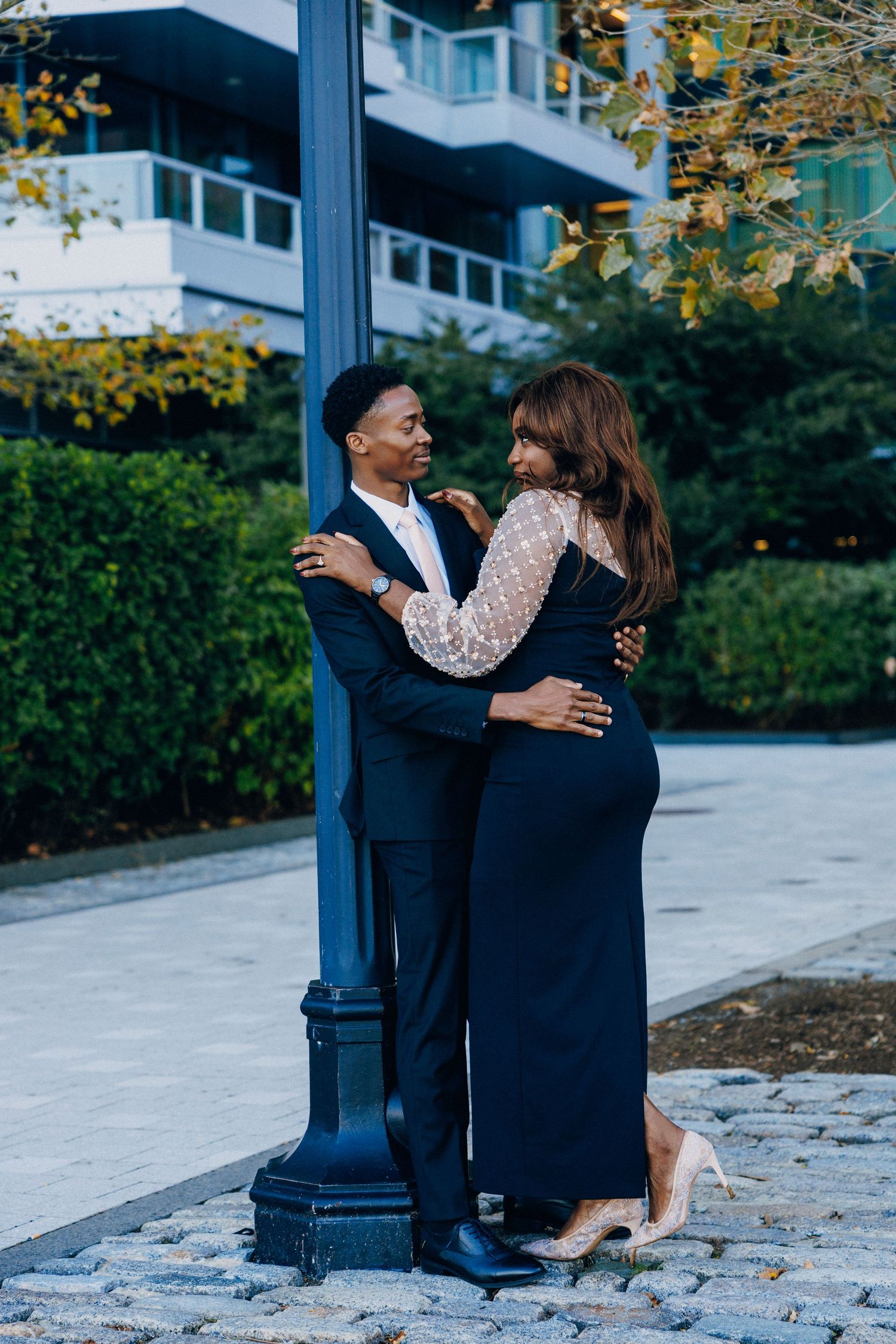 Couple in elegant formalwear embracing by a lamppost outdoors, man in navy suit, woman in black gown with sheer sleeves.