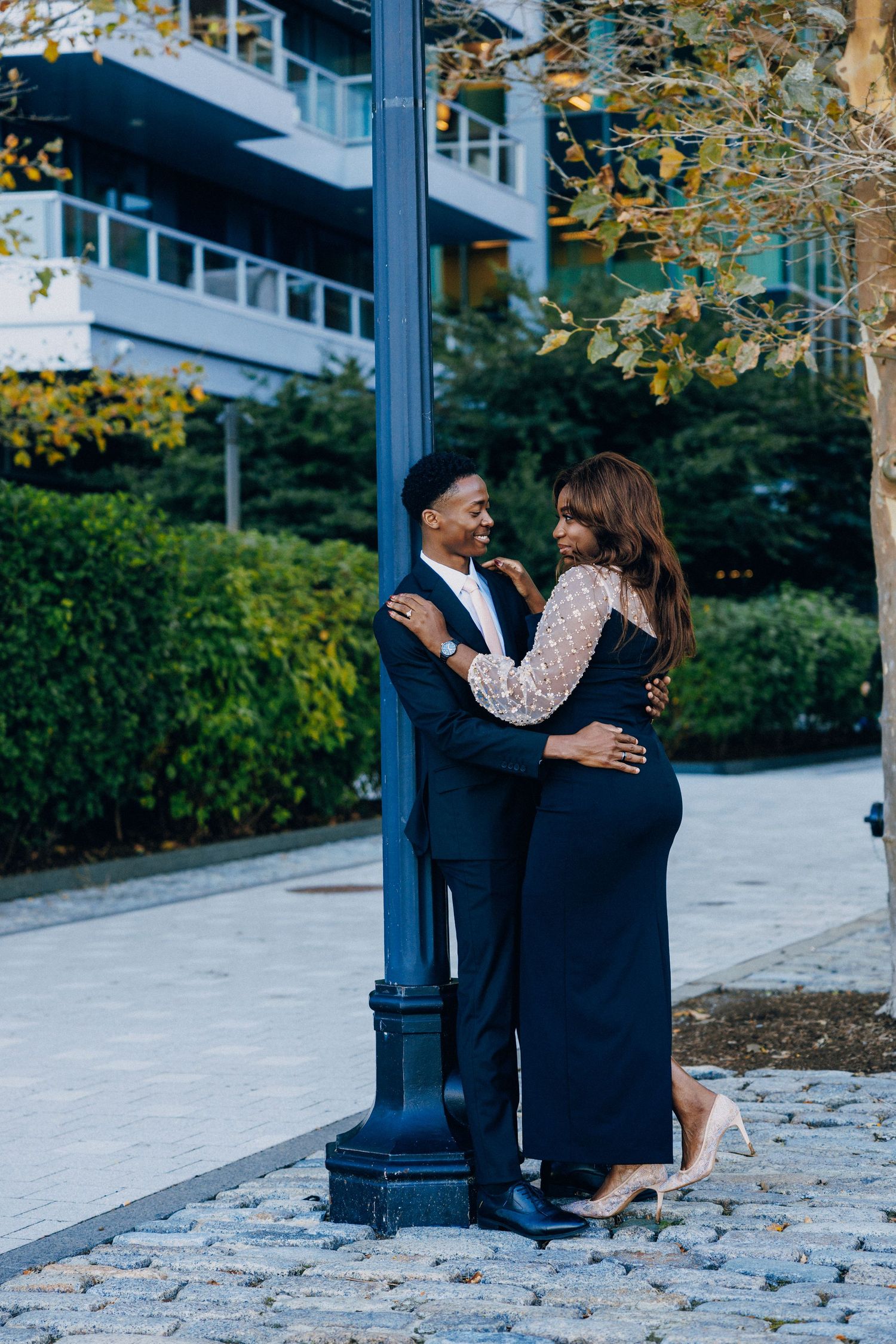 Elegant couple embracing by a blue lamppost on cobblestone path, wearing formal attire in an urban park setting.