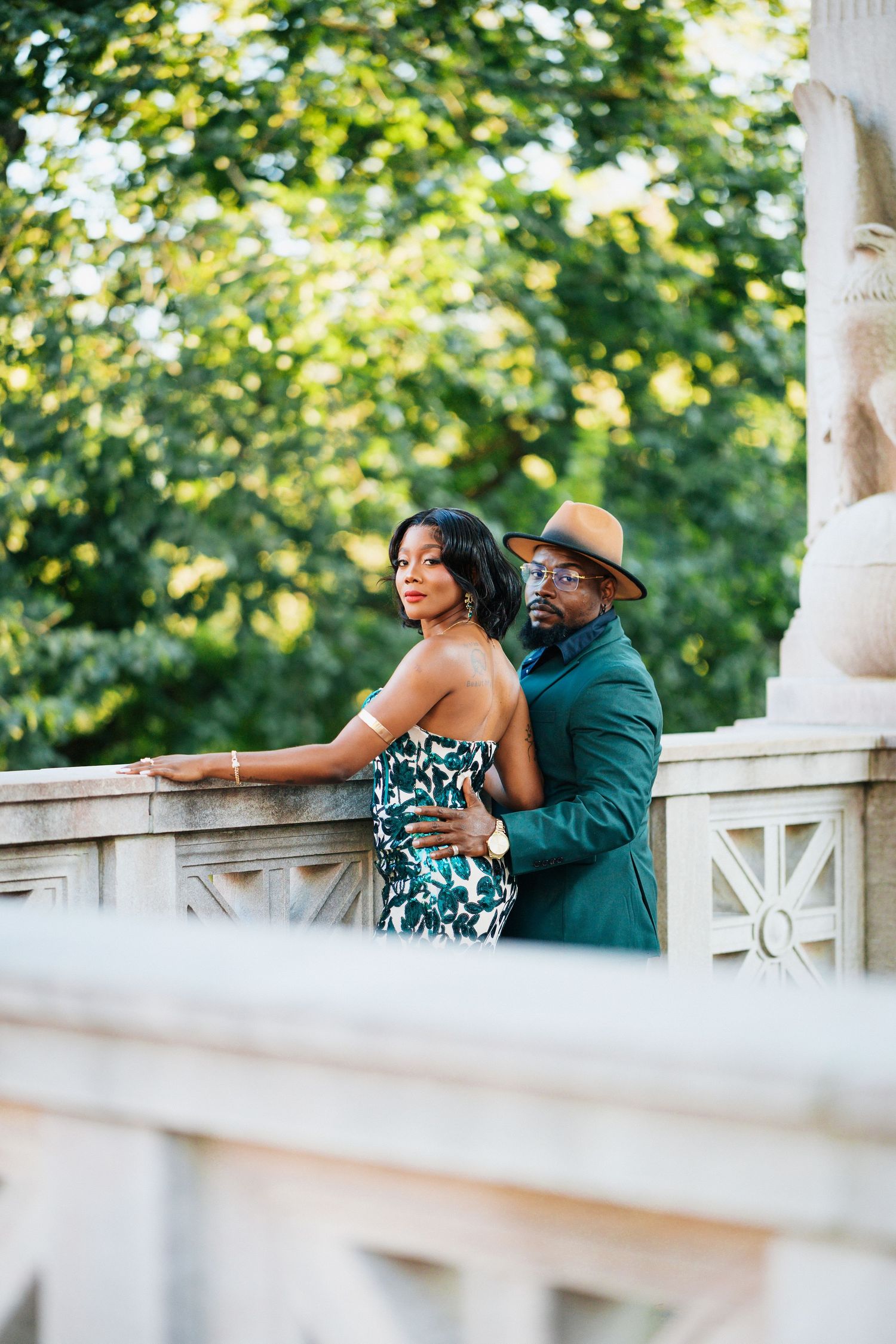 Elegant couple posing on a stone balcony surrounded by lush greenery, man in teal suit and hat embracing woman in floral gown.