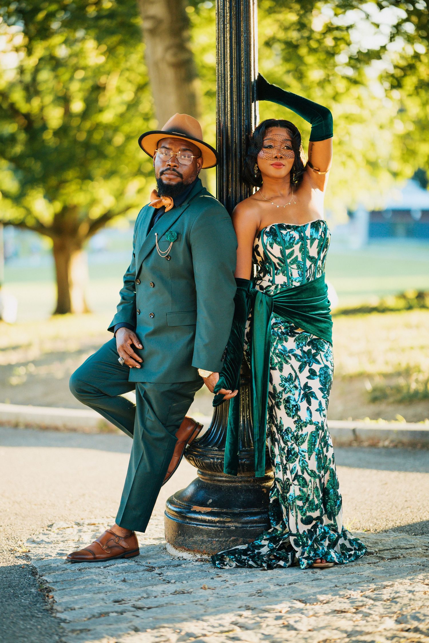 Fashionable couple in coordinated green outfits leaning against vintage lamppost in sunny park.
