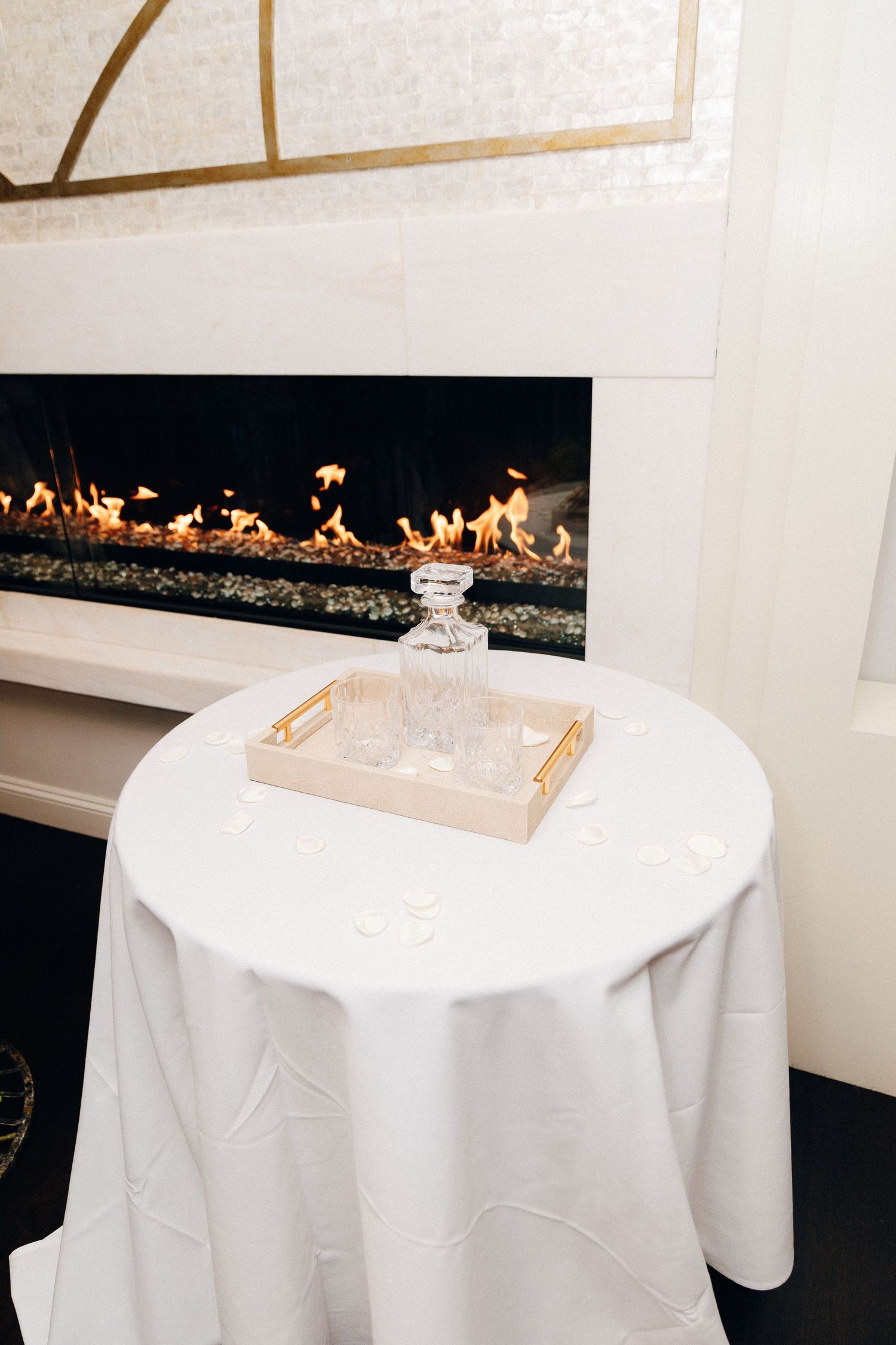Elegant white round table with crystal decanter on tray before modern linear fireplace.