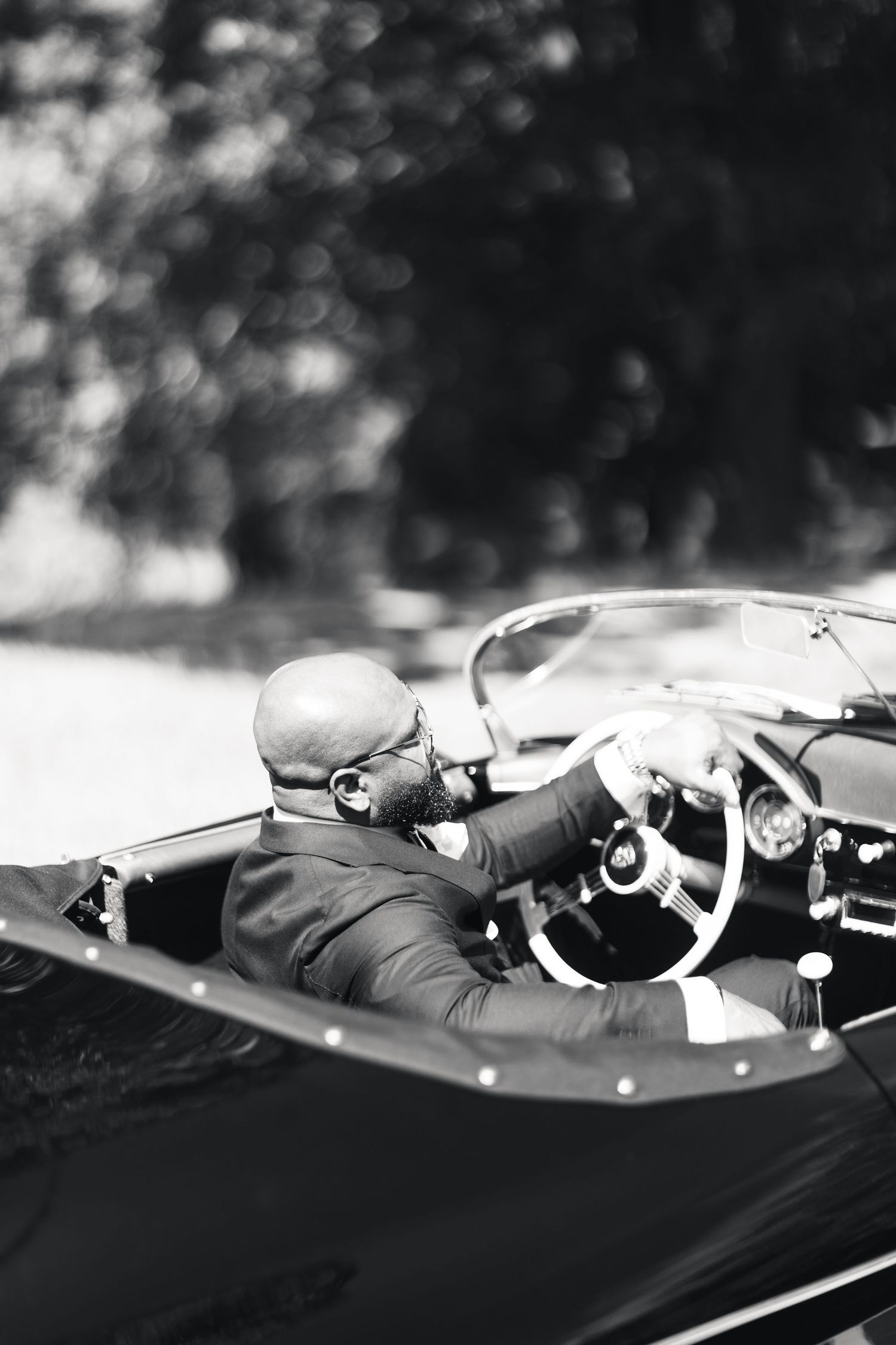 Black and white photo of groom driving a classic vintage convertible on his wedding day.