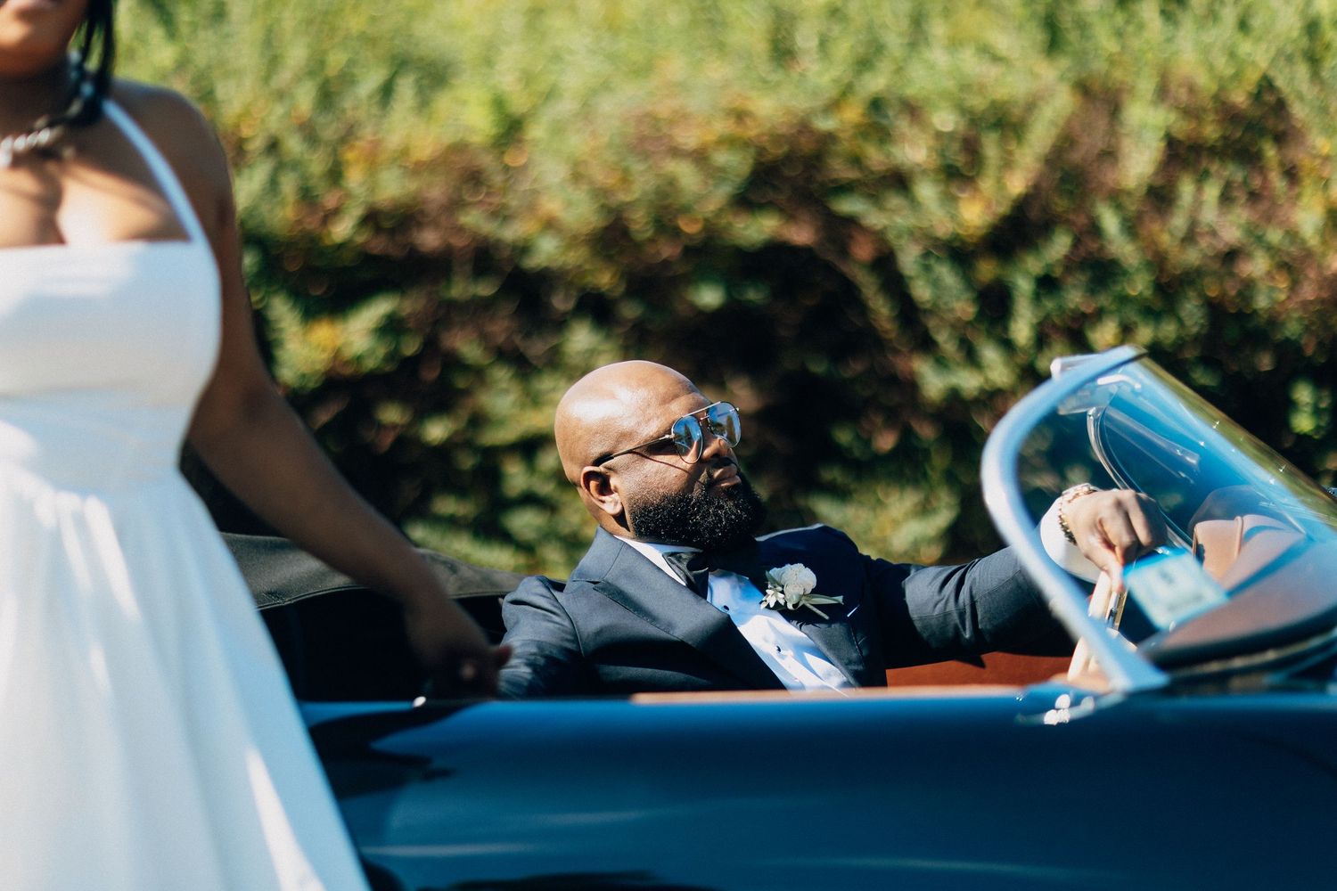 Groom in navy suit sits in a vintage convertible car wearing sunglasses on his wedding day.