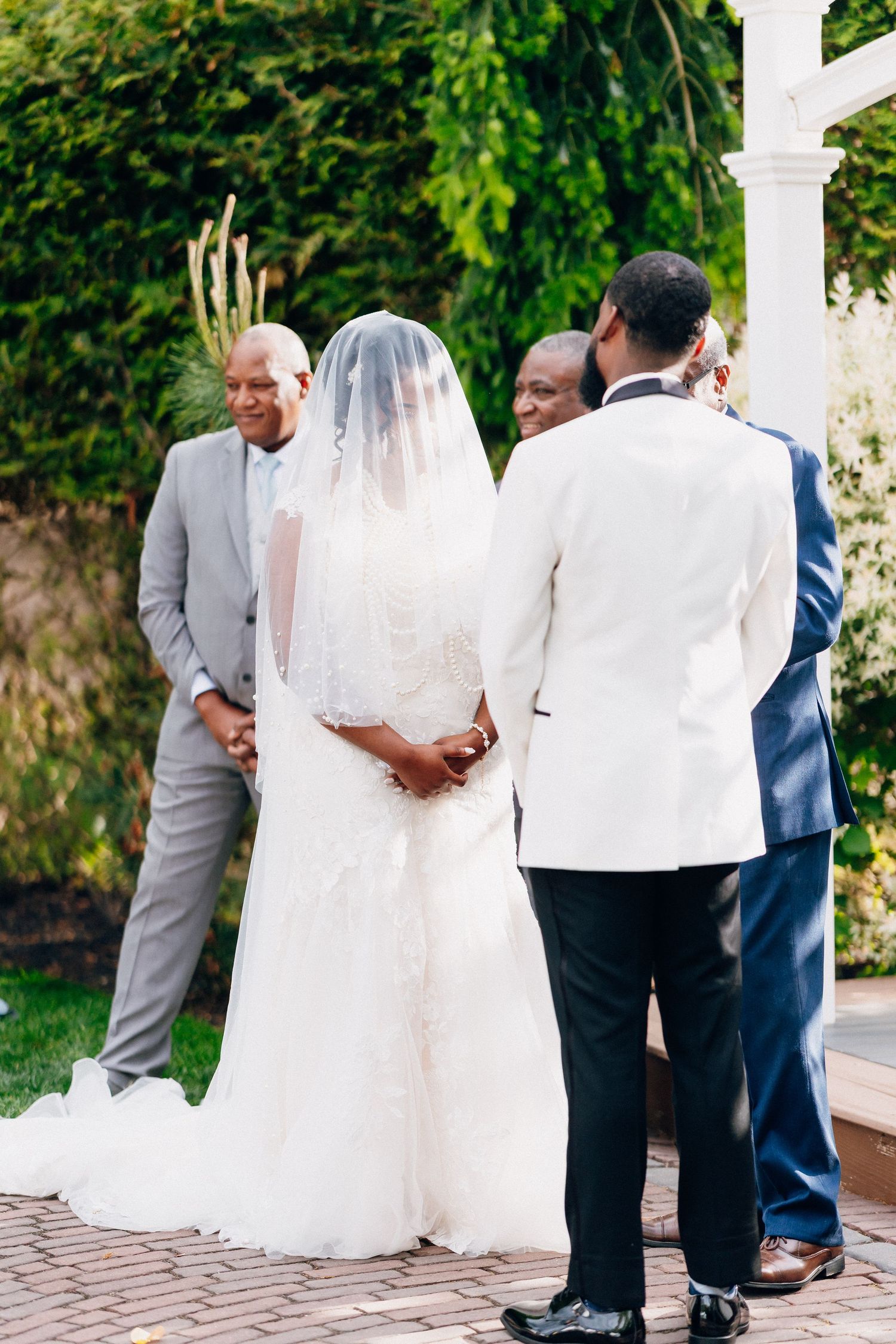 Bride in lace gown and veil stands at outdoor altar with groom in white jacket.