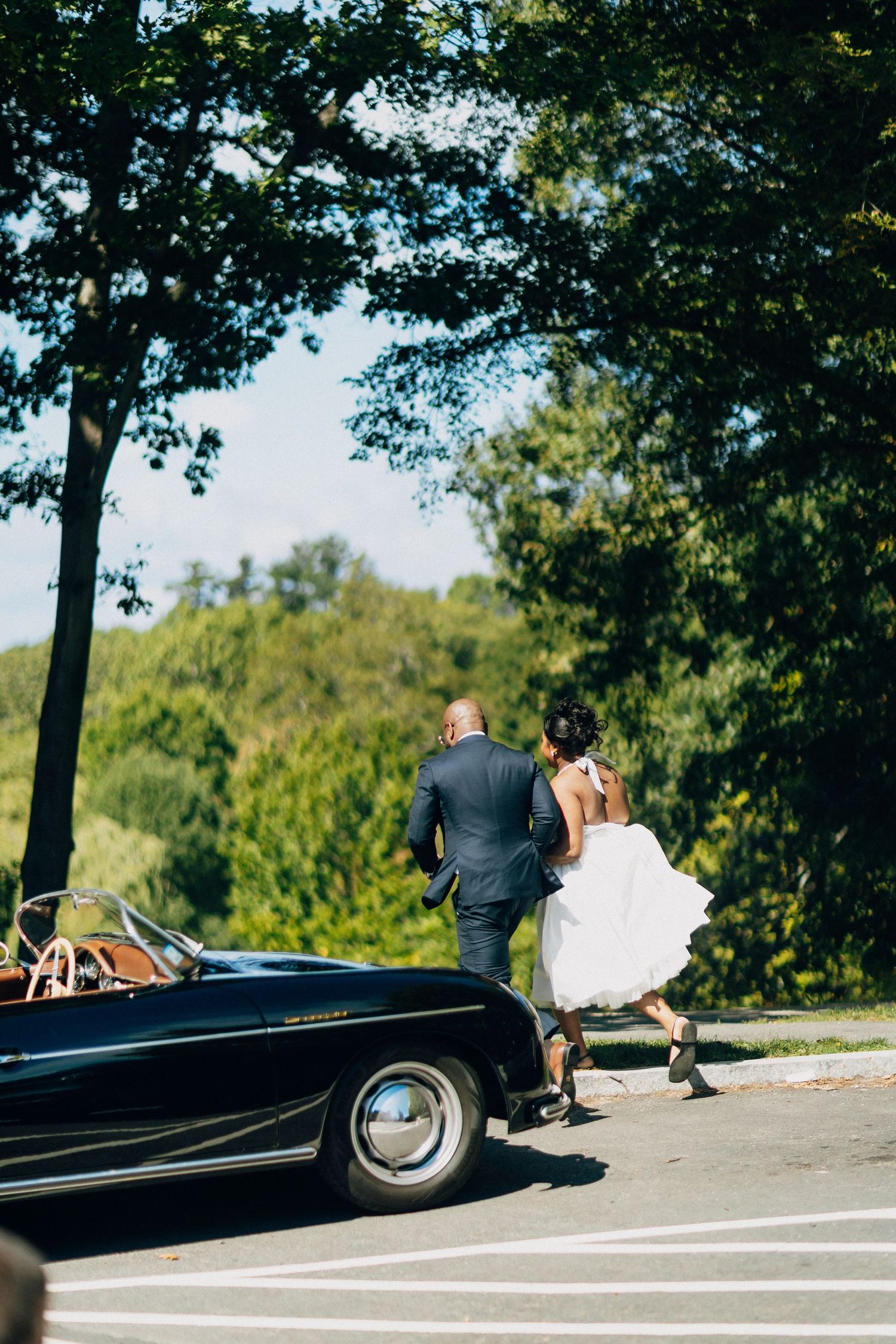 Bride and groom walk hand-in-hand toward a classic black convertible car on their wedding day.