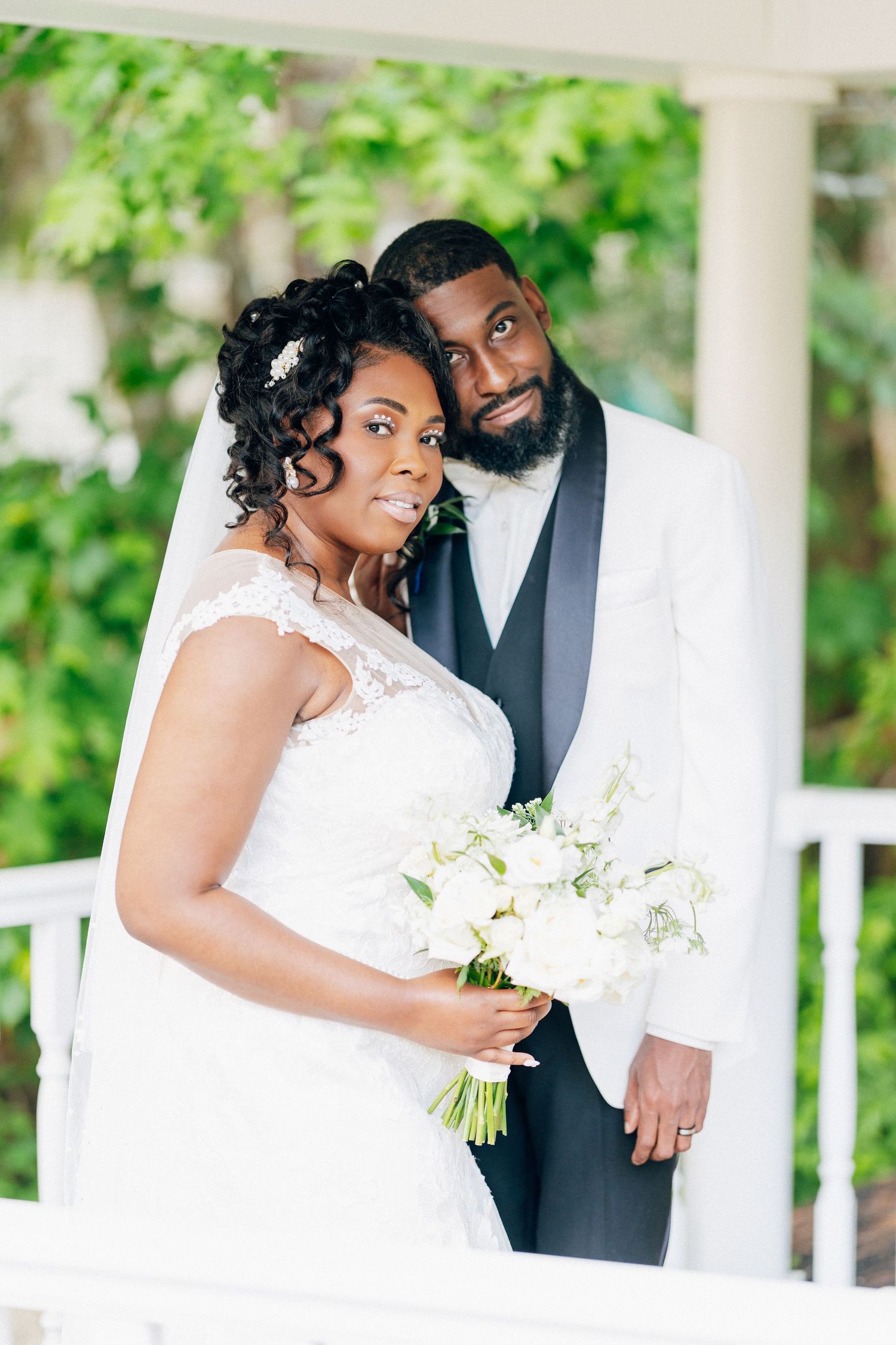 A bride and groom pose elegantly on a white gazebo, she holds white flowers in a lace gown, he wears a white tuxedo.