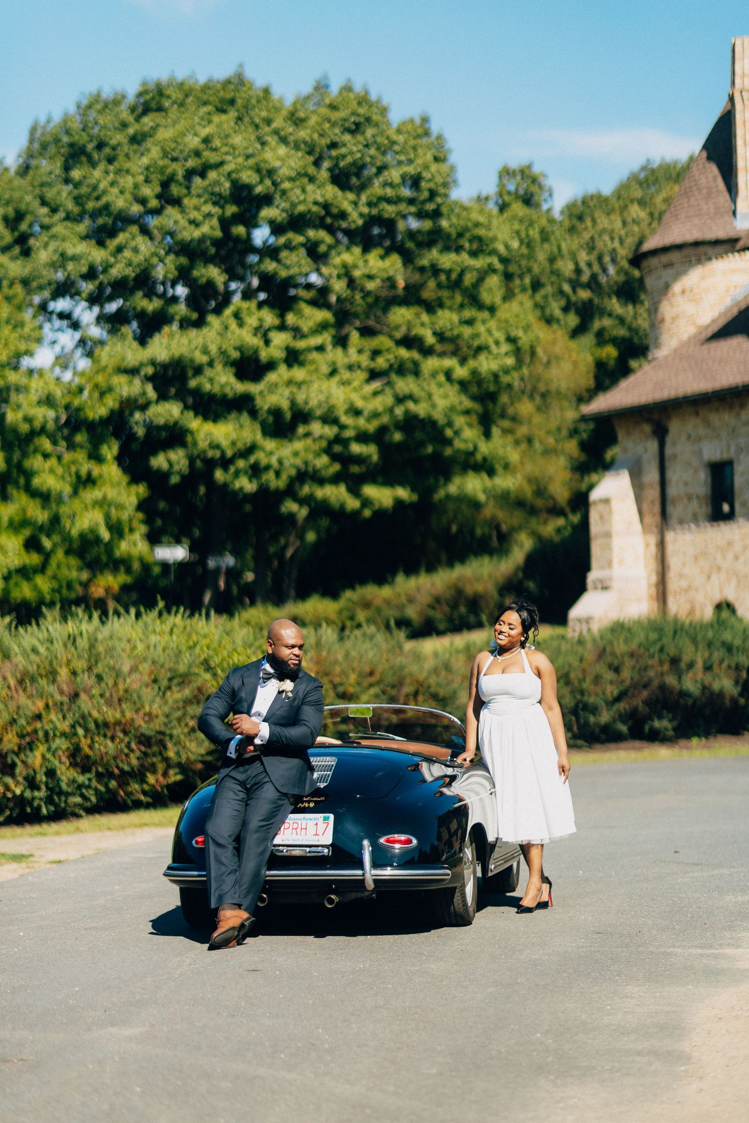 Elegant couple in wedding attire pose beside vintage black Porsche 356 convertible near stone building.