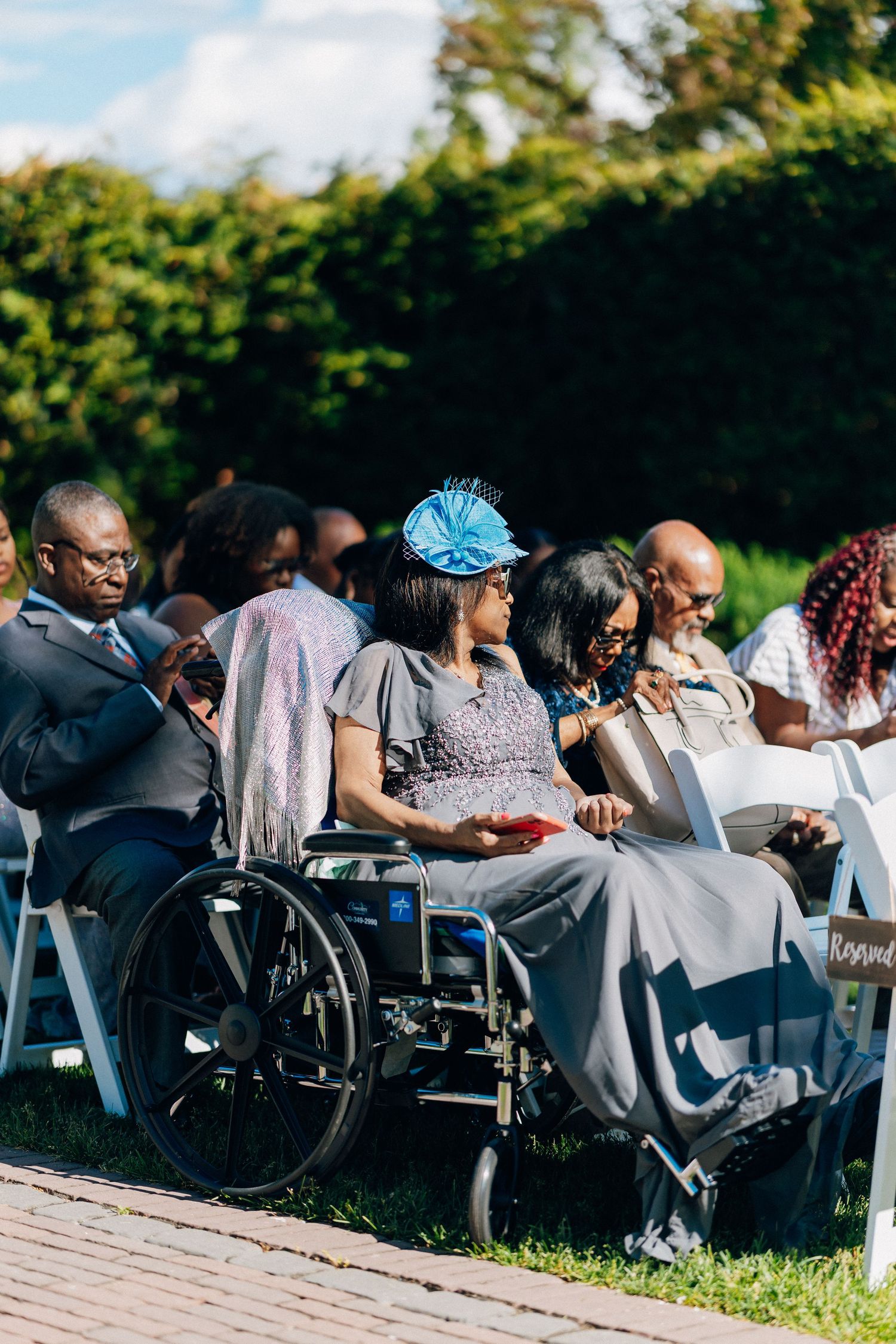 Guests seated at an outdoor wedding ceremony, including a woman in a blue fascinator hat in a wheelchair.