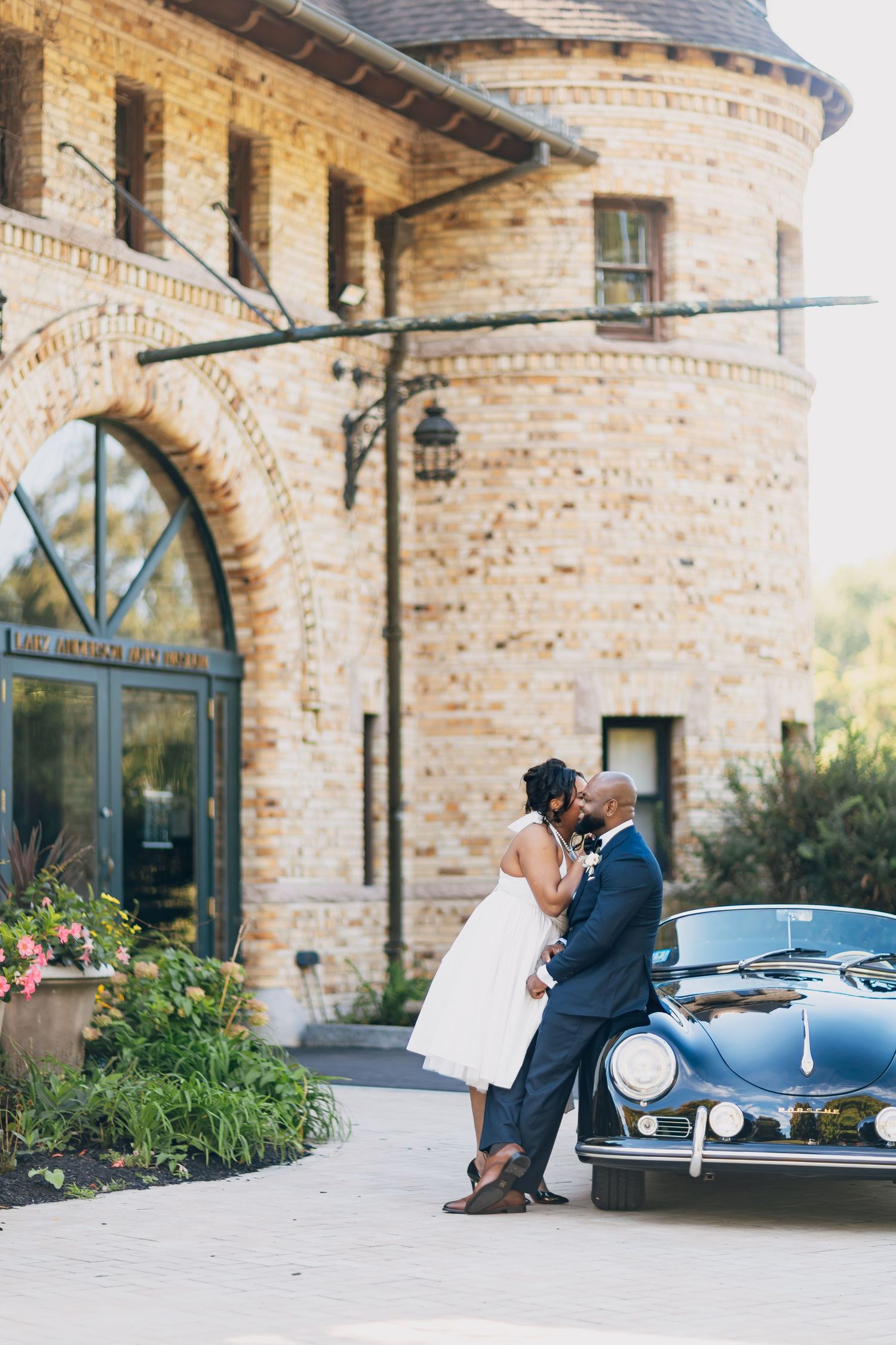 Bride and groom share a romantic kiss beside a vintage Porsche in front of a stone castle venue.