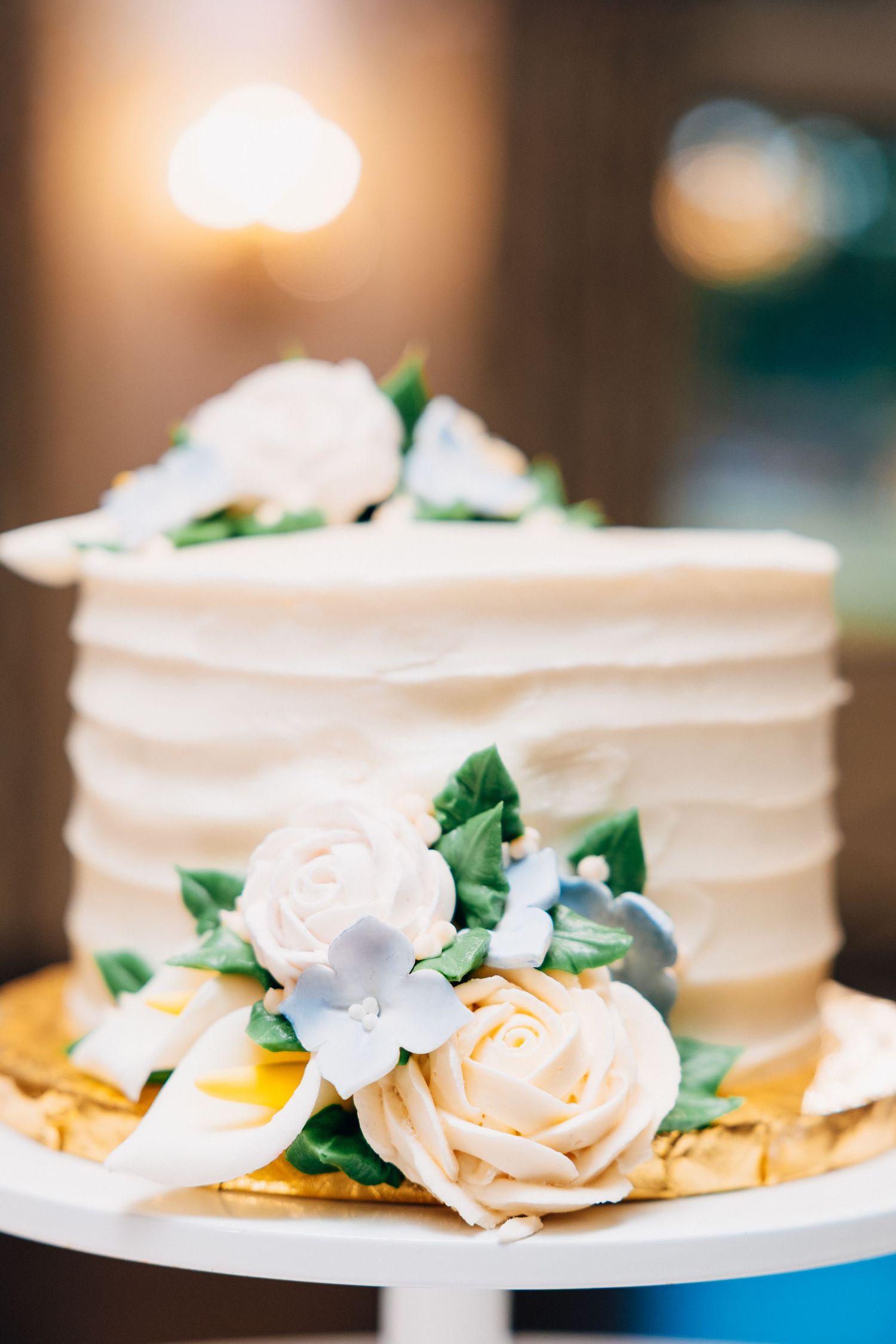 Elegant two-tier white wedding cake decorated with cream and blue sugar flowers on a white stand.