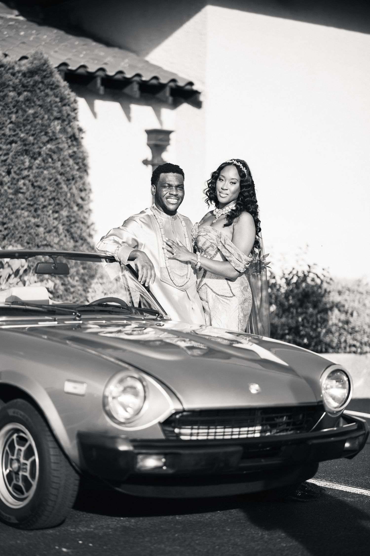 Couple in traditional African attire posing beside a classic vintage convertible car in black and white photo.
