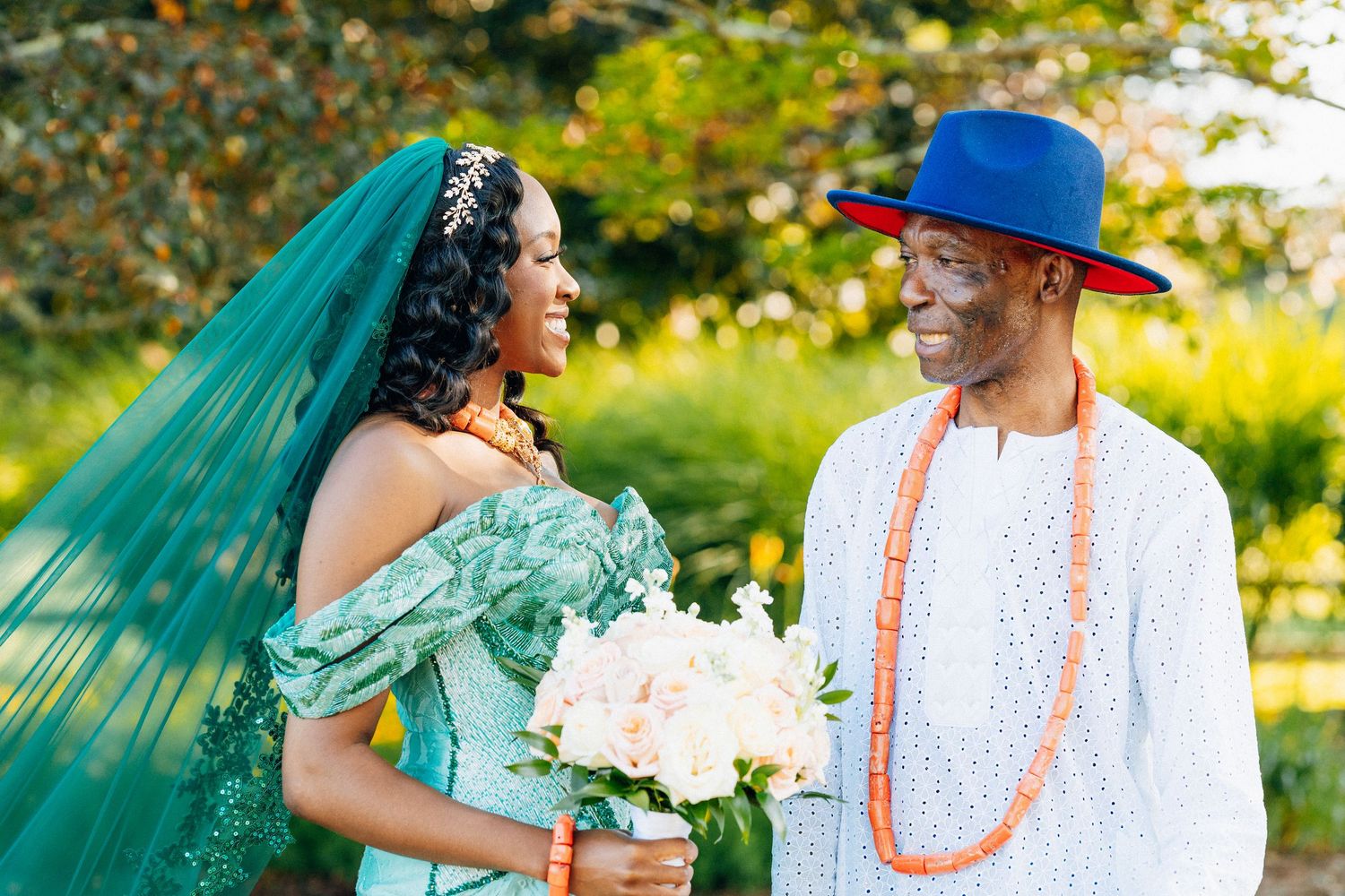 Couple in Nigerian traditional wedding attire, bride in teal gown with veil, groom in white with orange beads and blue hat.