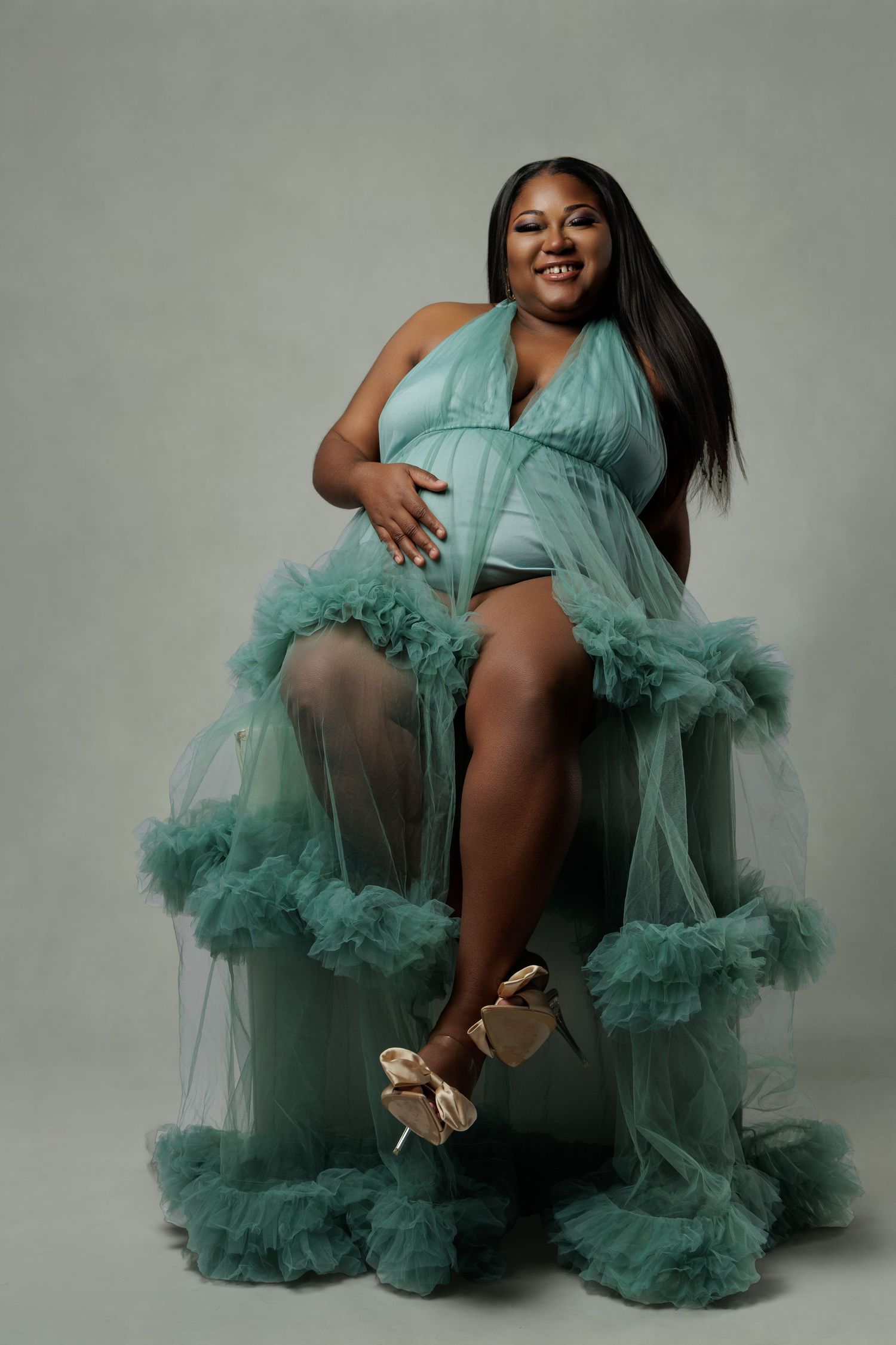 Pregnant woman in teal ruffle tulle maternity gown sitting gracefully, wearing gold heels against neutral background.
