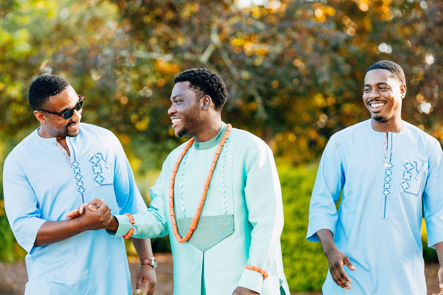 Three men in matching light blue African traditional attire laughing and celebrating outdoors.