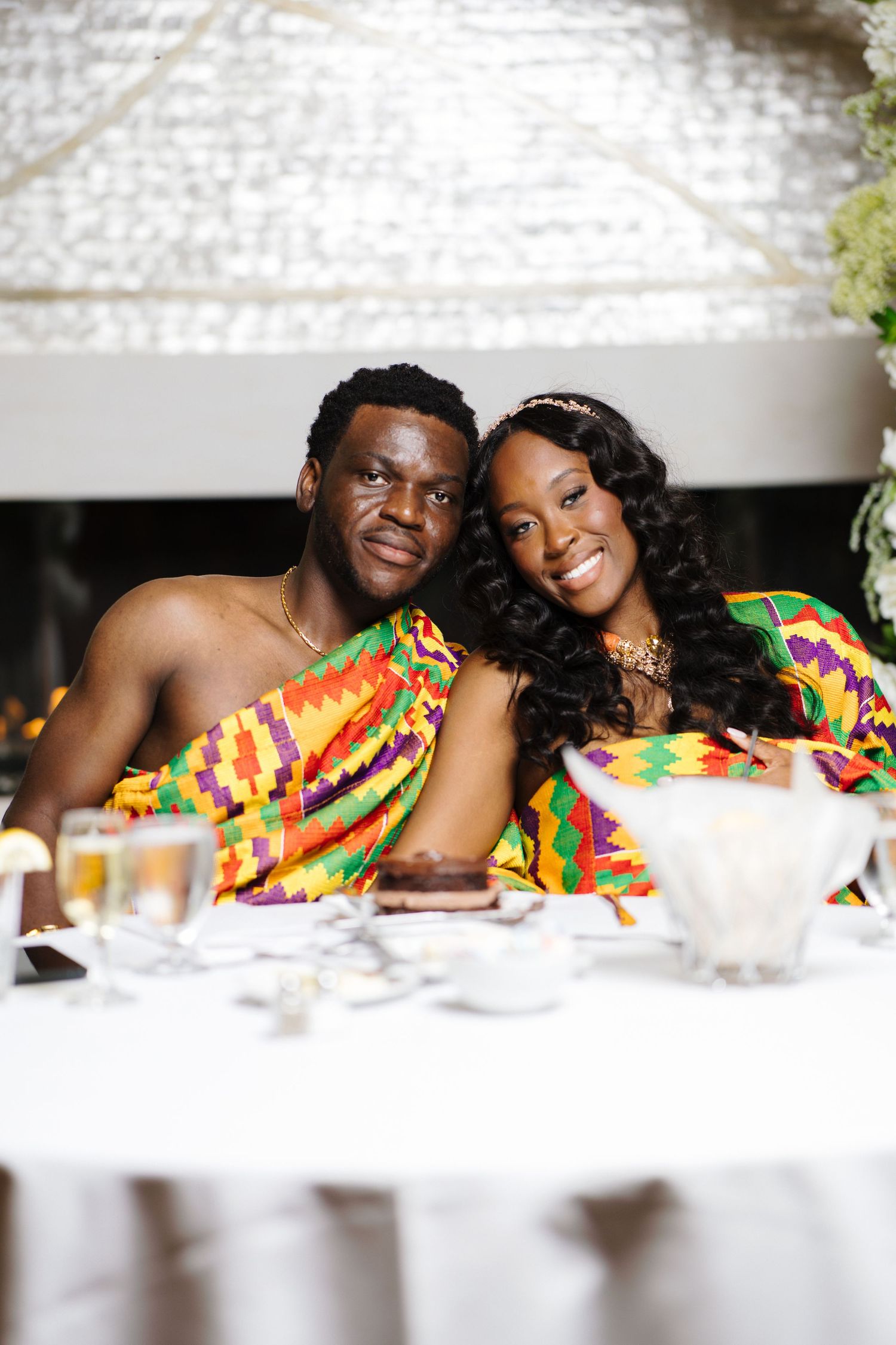 Smiling newlywed couple in traditional Kente attire seated together at their wedding reception table.