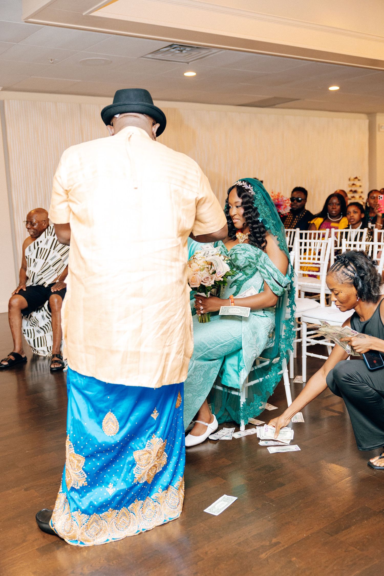Man in gold African attire presents bride in teal gown during traditional wedding ceremony.
