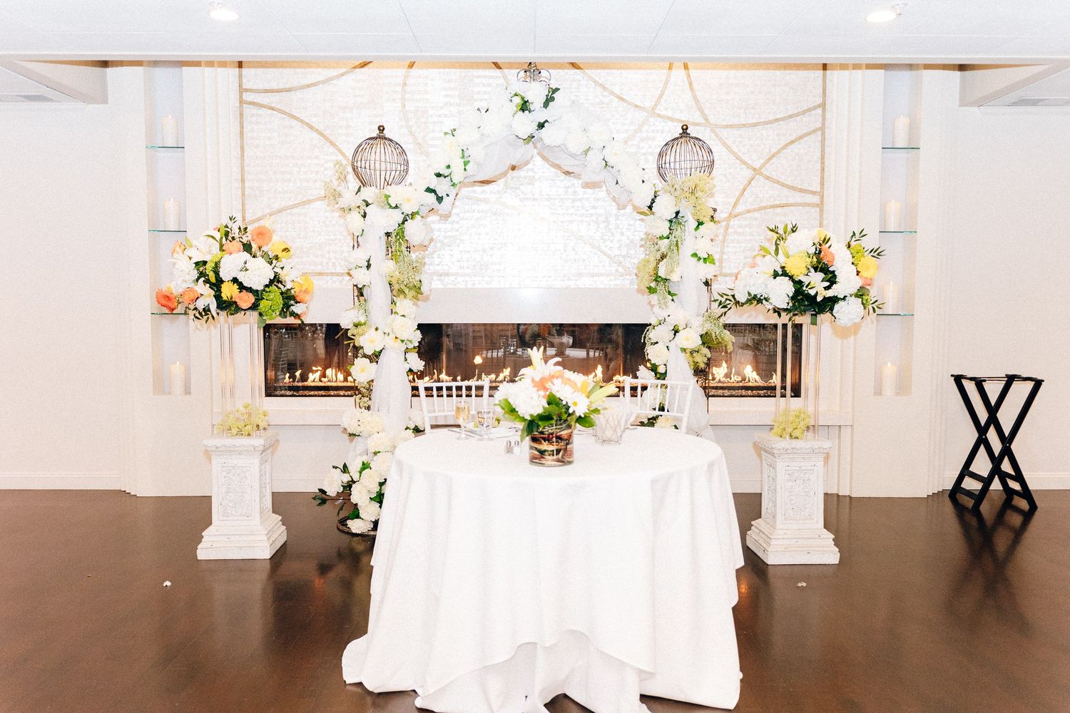 Elegant wedding sweetheart table with white floral arch, birdcages, and fireplace backdrop.