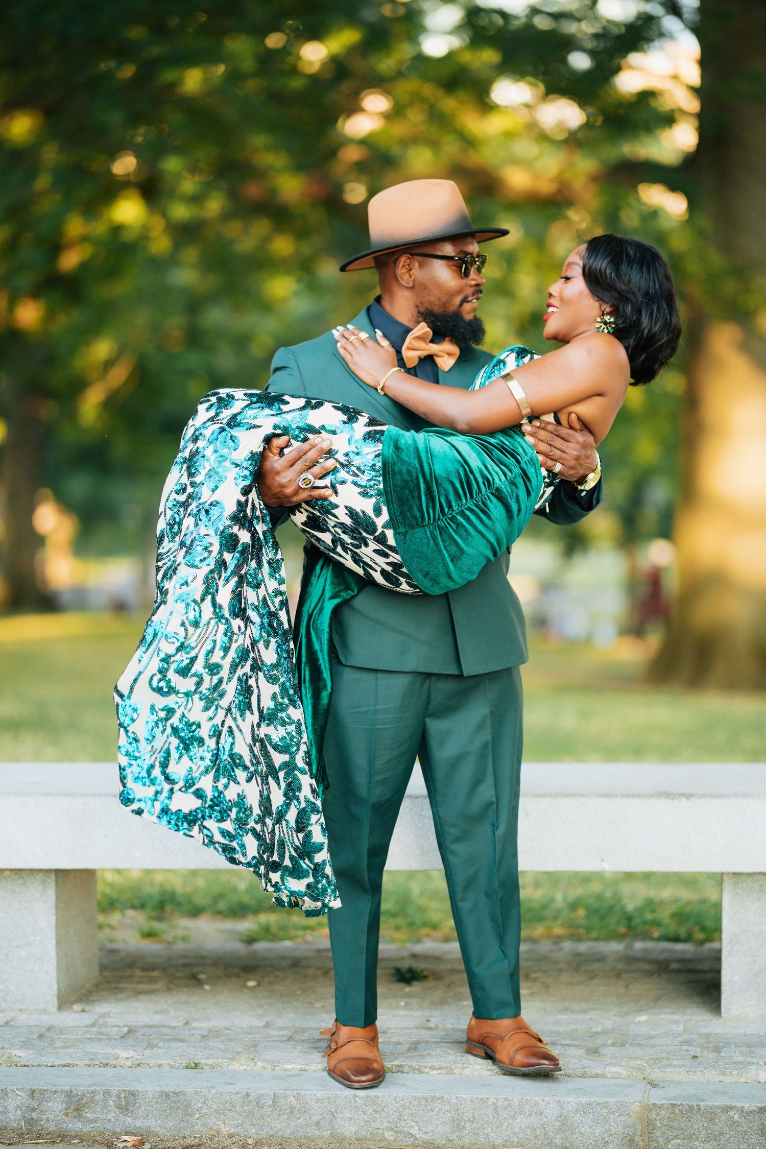 Dapper Black man in green suit lifting woman in floral gown in sunlit park.