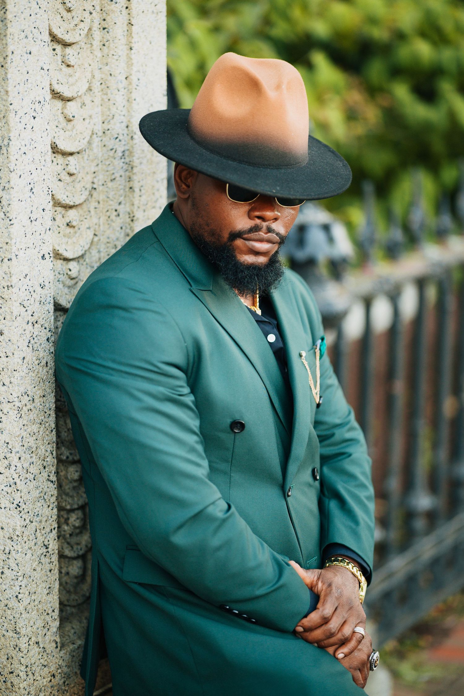 Dapper man in emerald green double-breasted suit and tan fedora leaning against stone column.
