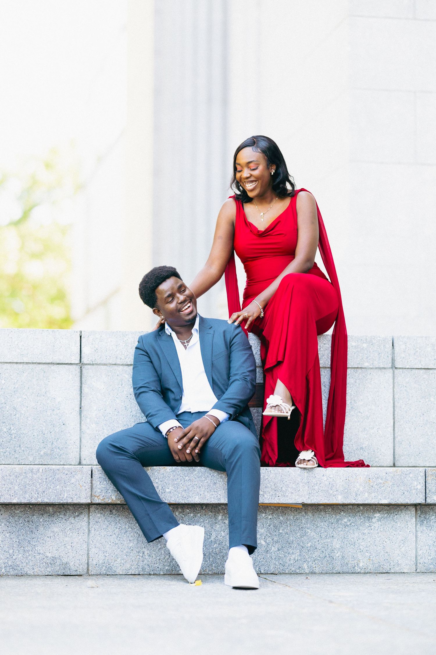 Joyful couple on marble steps, man in gray suit seated while woman in red dress smiles.