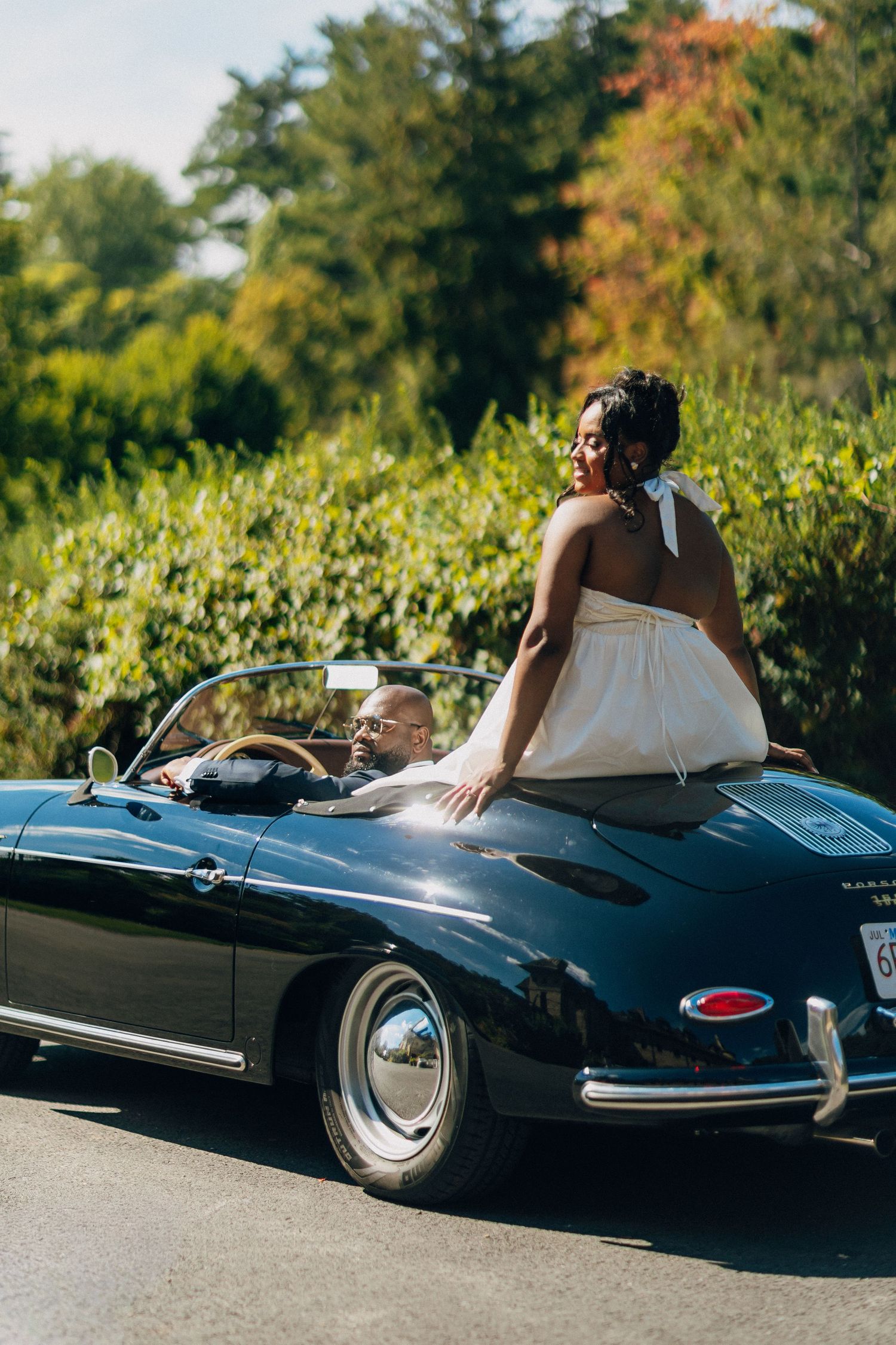 Joyful Black bride sits on the back of a vintage black convertible Porsche driven by her groom.