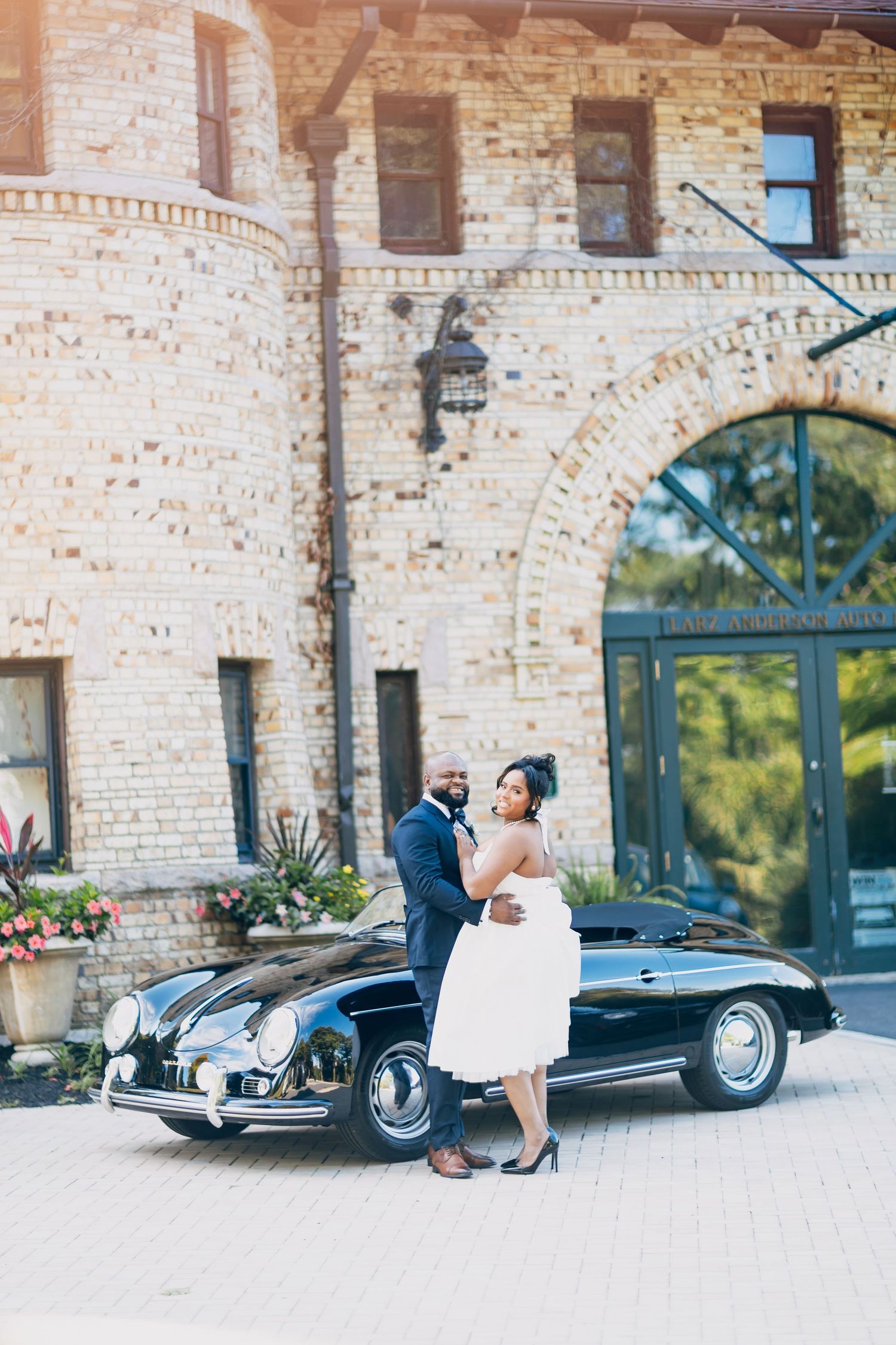 Elegant bride and groom posing beside a classic black Porsche outside a stunning brick building.