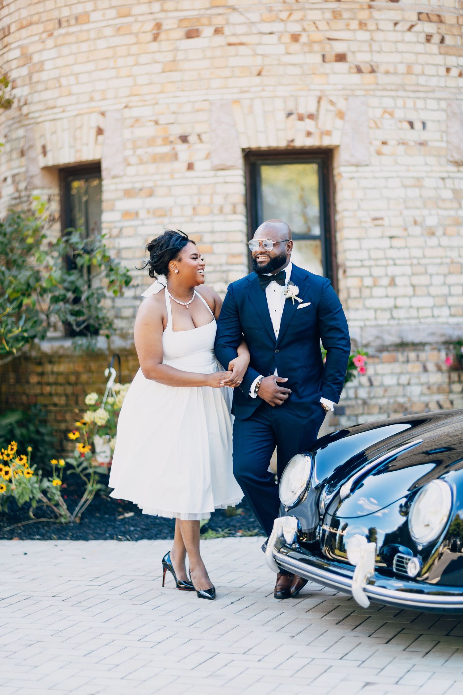 Black bride and groom walking arm in arm beside a vintage car outside a brick building on their wedding day.