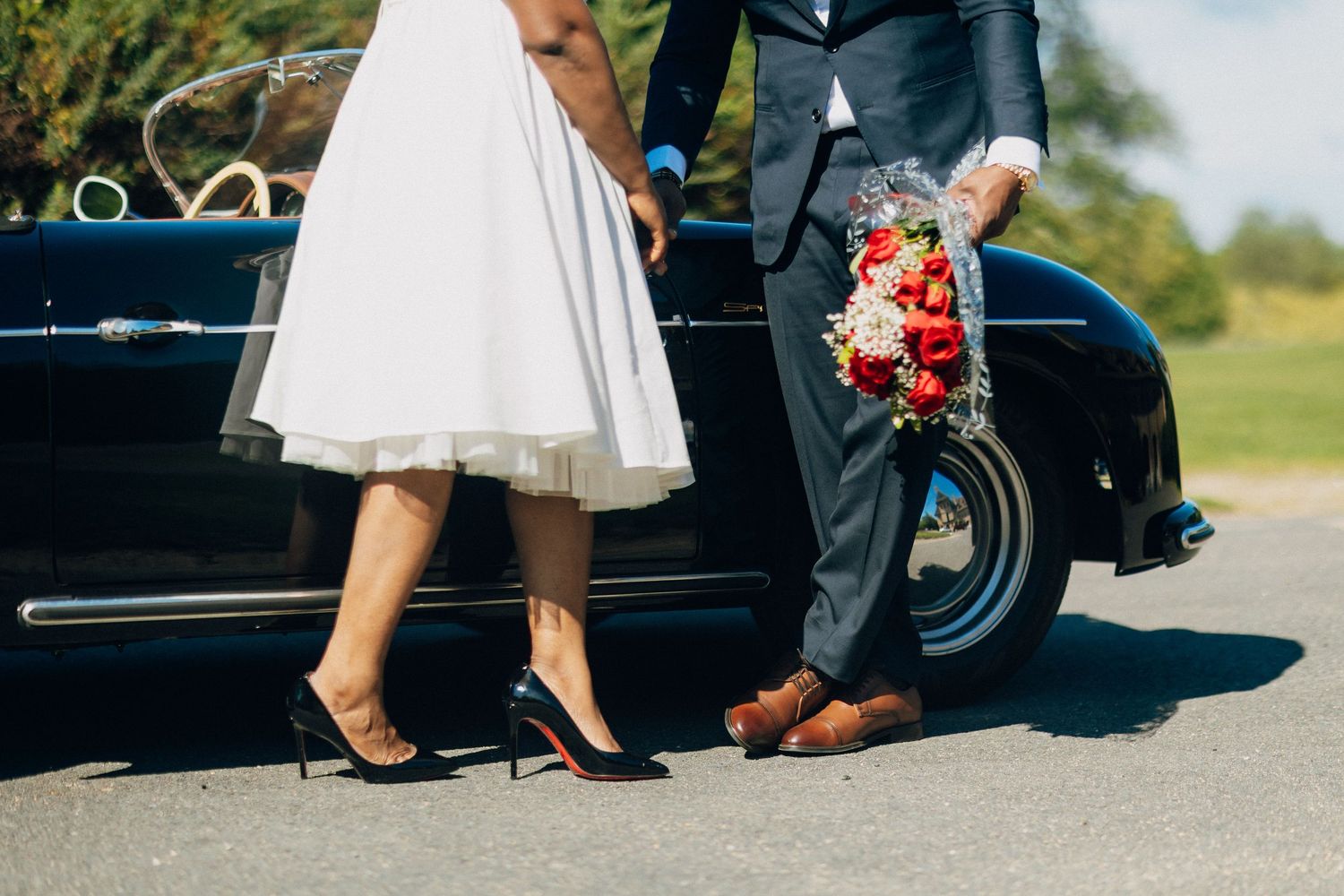 Close-up of bride in heels and groom holding red bouquet standing beside a vintage black car.
