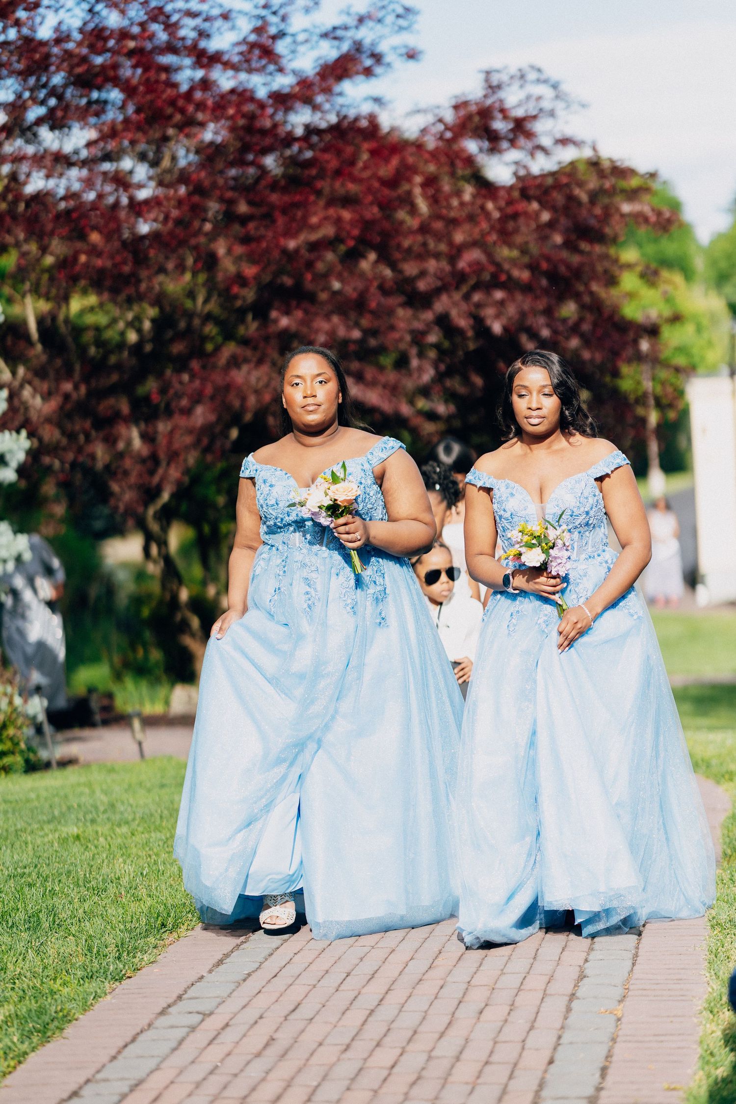 Two bridesmaids in powder blue off-shoulder ball gowns carrying bouquets walking down outdoor aisle.