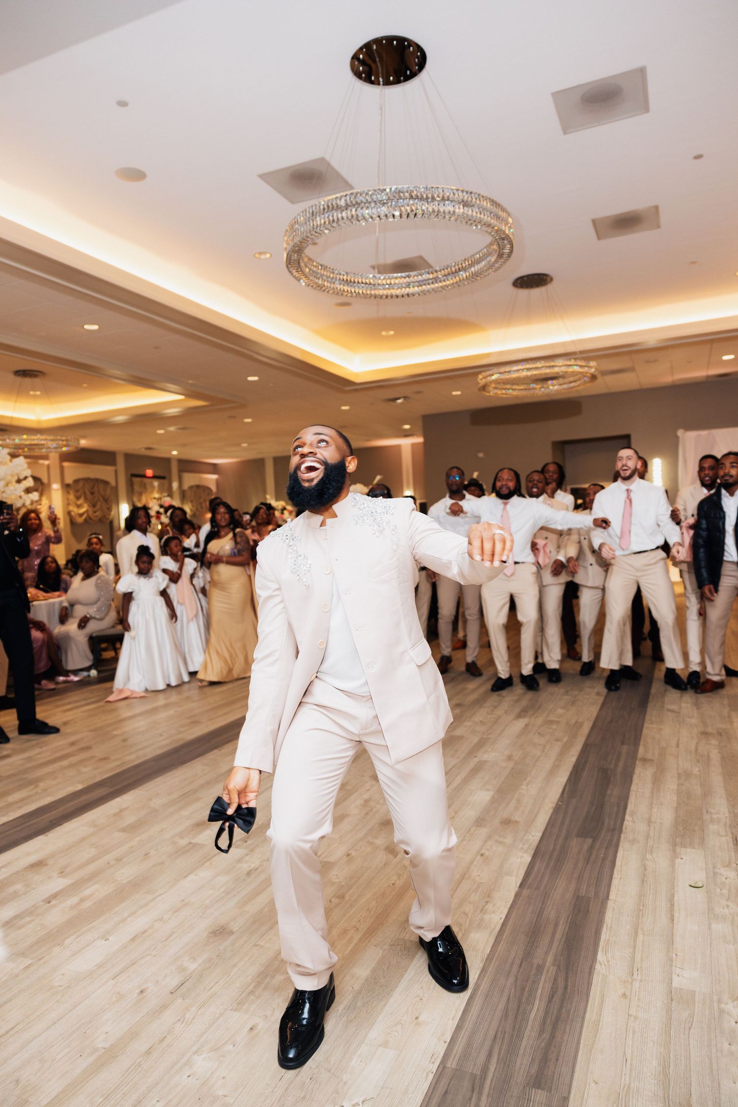 A joyful groom in a white suit dances energetically on the wedding reception dance floor while guests watch in the background.