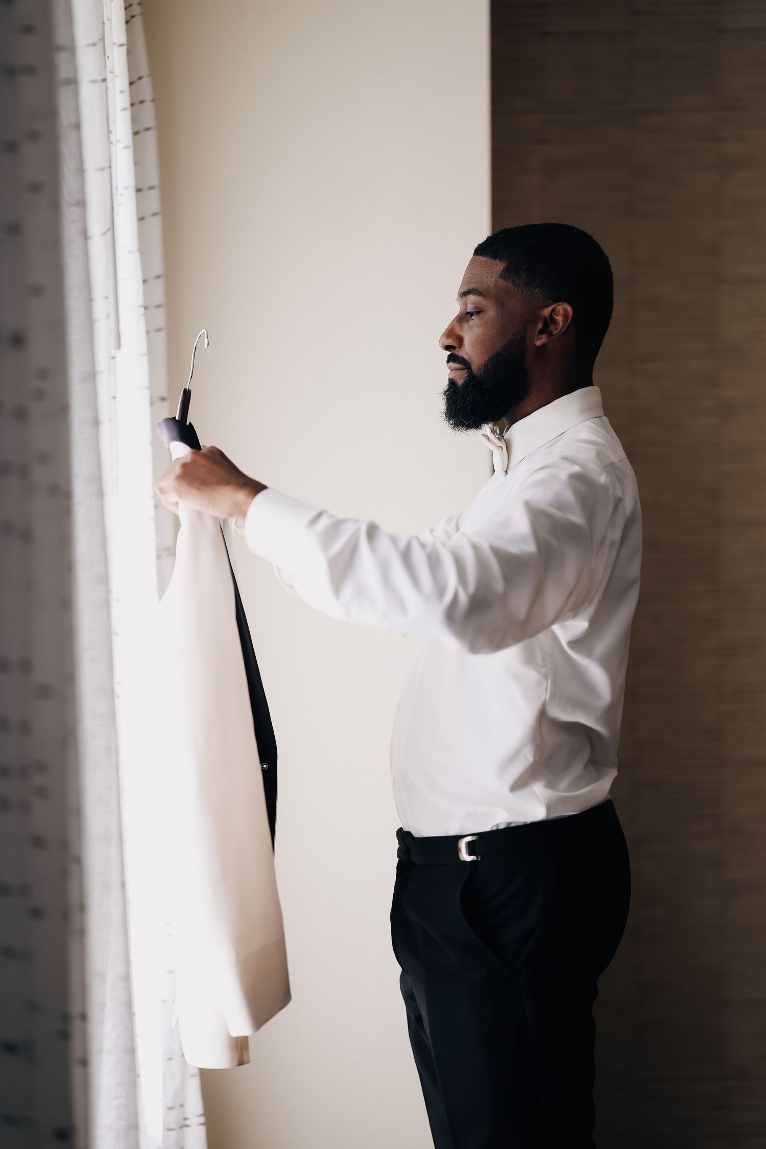 Bearded Black groom in white dress shirt examining suit jacket on hanger near window.