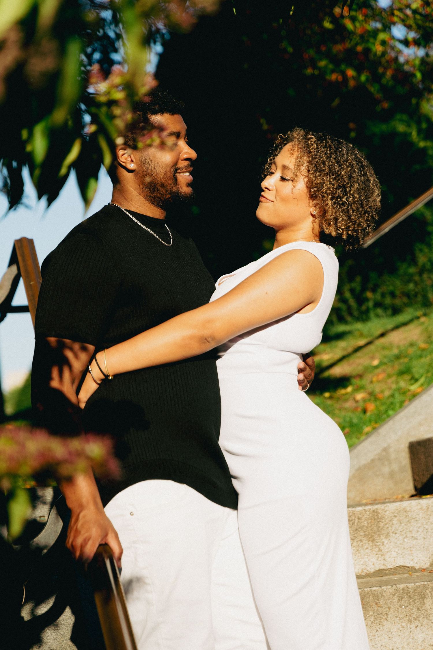 A couple embraces on outdoor steps, woman in white dress and man in black shirt, surrounded by lush greenery.